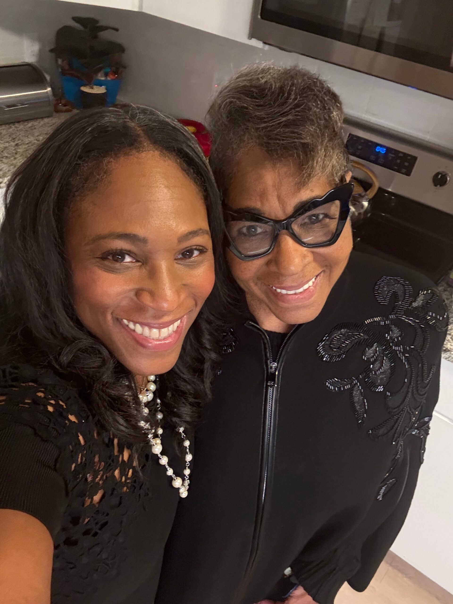 Two women smiling for a selfie in a kitchen, one in a lace black top with pearls and the other wearing glasses and a black embroidered jacket.