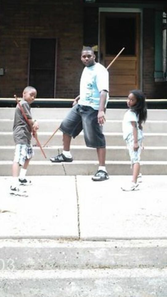 A man stands on steps with two children, each holding a wooden stick as if ready to practice martial arts in a casual outdoor scene.