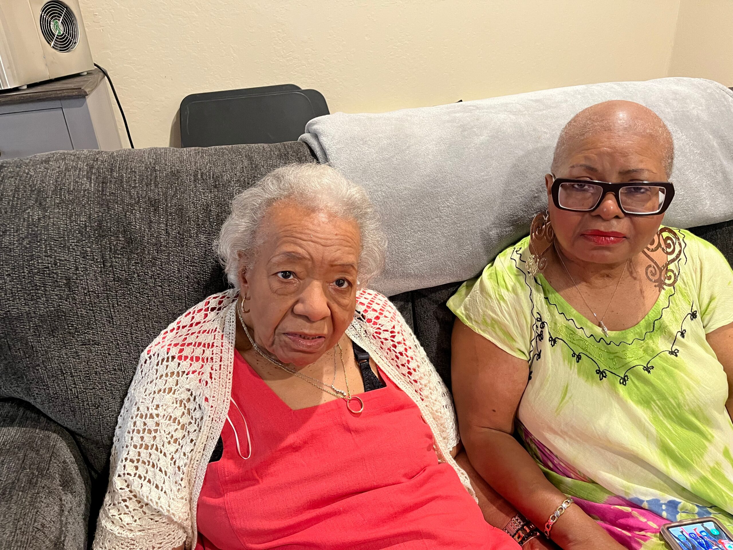 Two smiling women sit close together on a gray couch, wearing bright red and lime outfits, in a cozy living room.