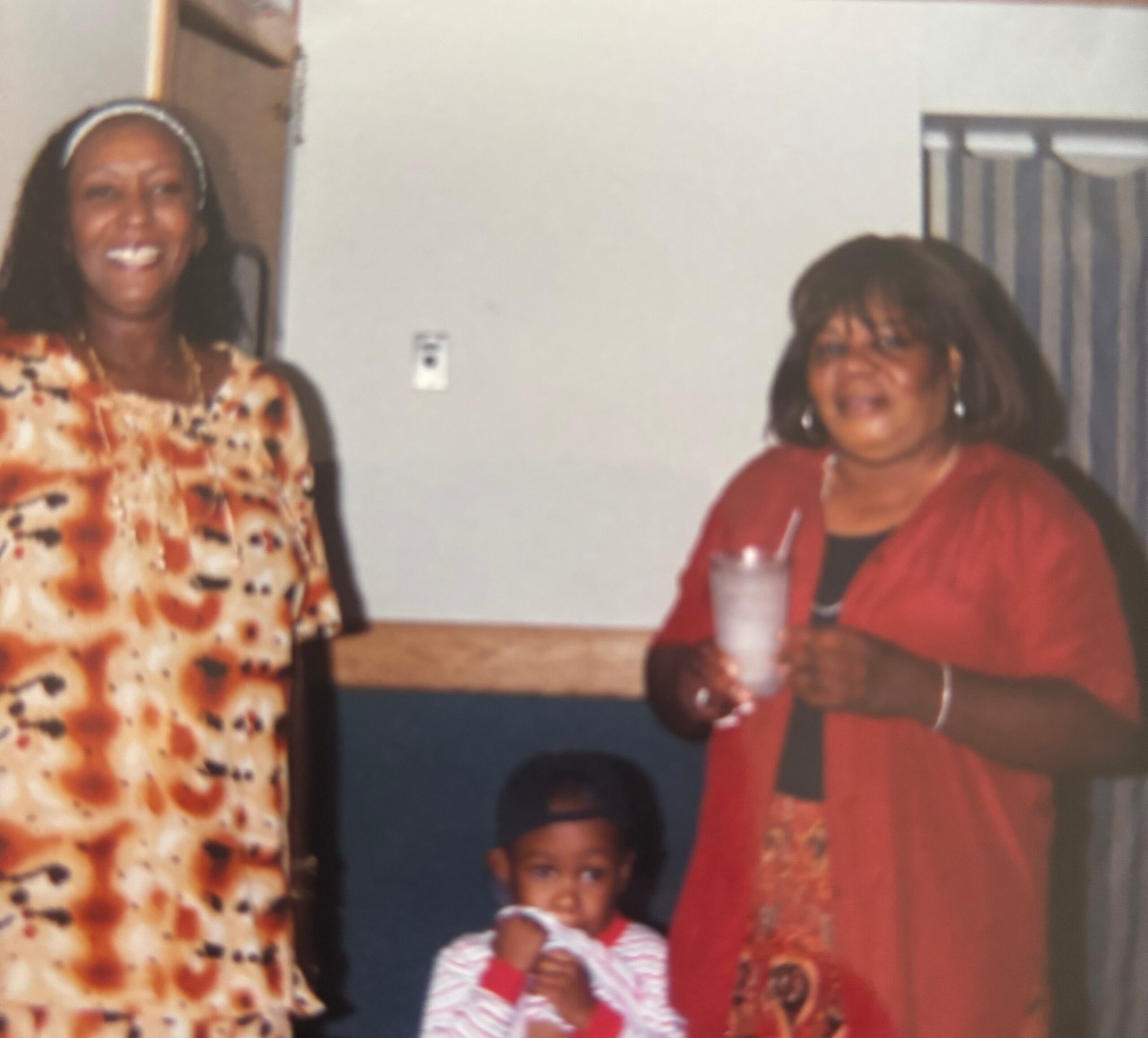 Two women posing indoors with two children; left woman in a floral dress, right woman in a red jacket, child in striped shirt seated at front.