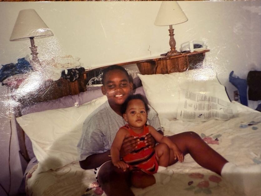 Man sitting on a bed with a baby on his lap, both smiling, in a bedroom with two lamps on the headboard.