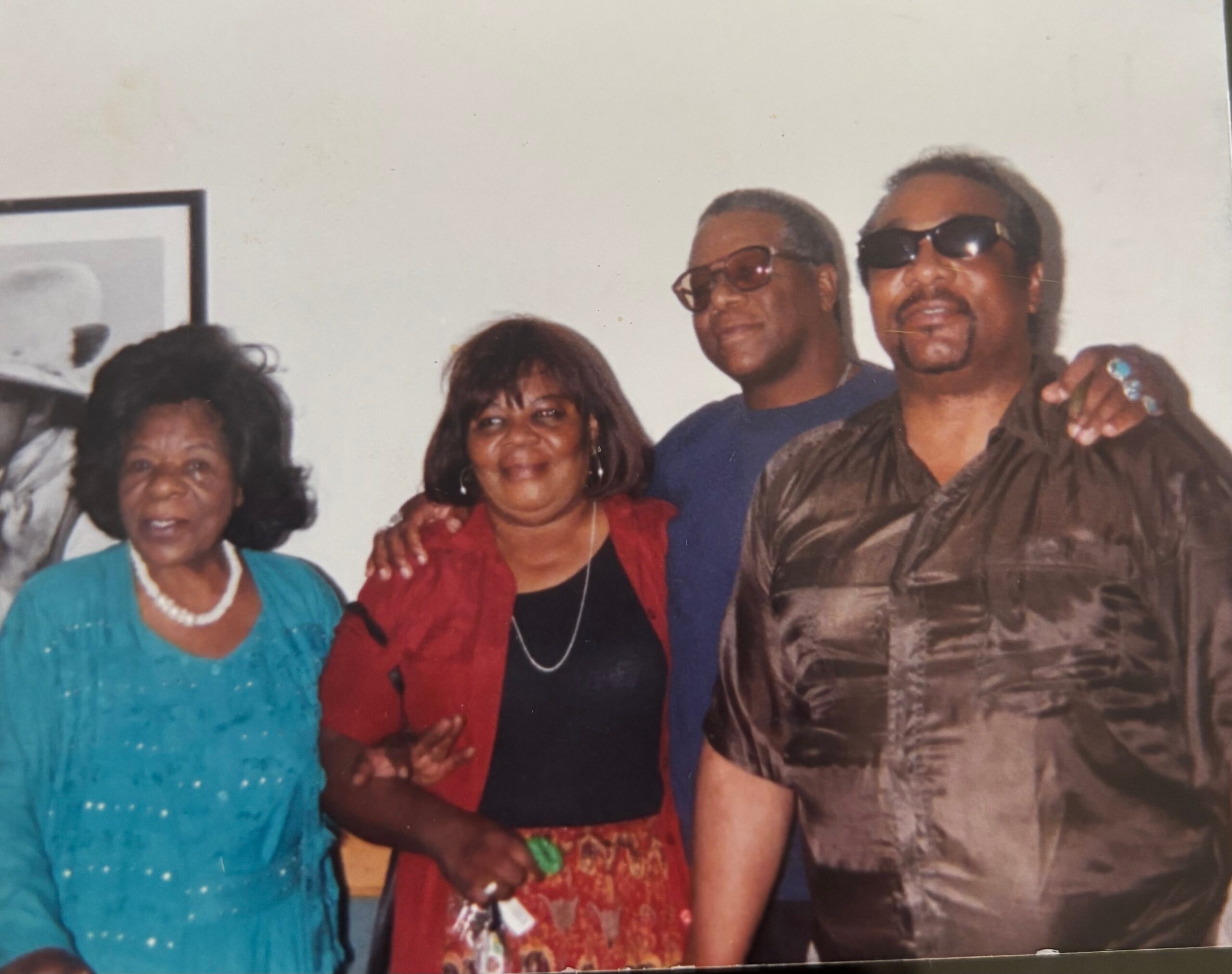 Four people posing together indoors, two women on the left wearing teal and red outfits, two men in sunglasses on the right.