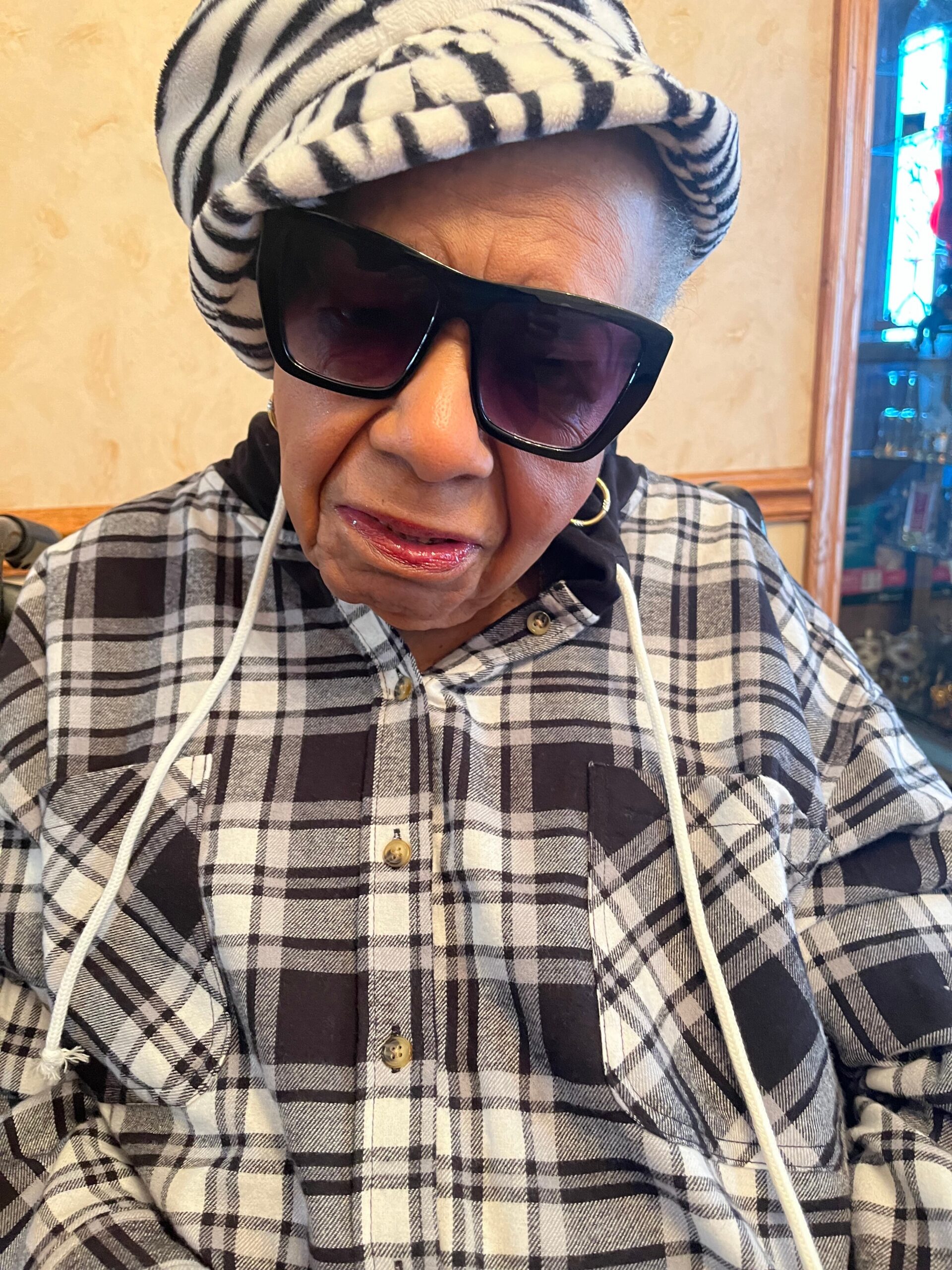 Older person wearing oversized black sunglasses and a zebra-print hat, with a black-and-white plaid shirt indoors.