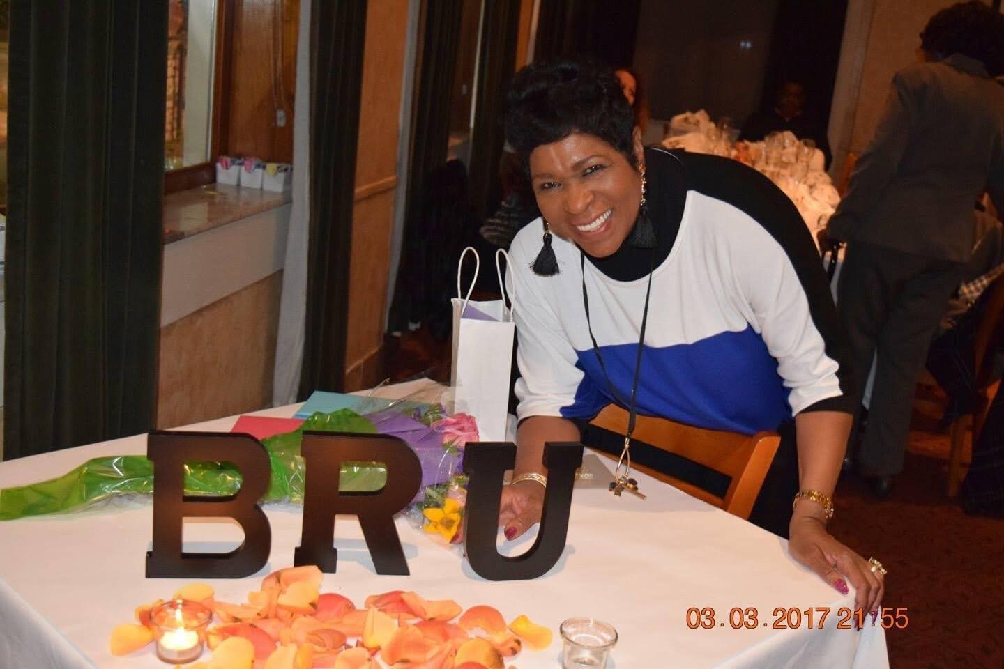 Smiling woman in a white and blue top leans over a decorated table with large black letters spelling BRU, surrounded by rose petals and candles at a banquet event.