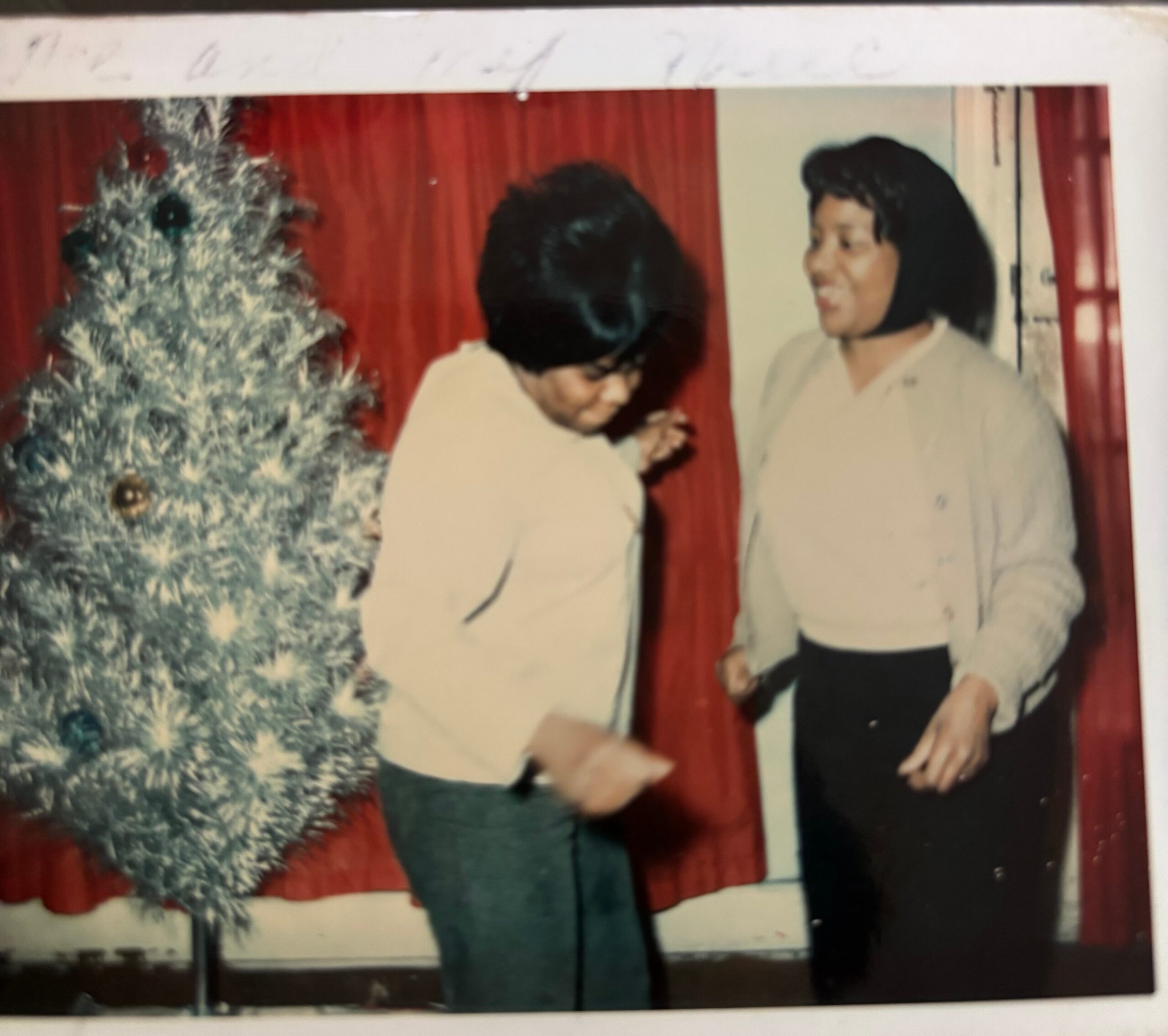 Two people stand near a frosted Christmas tree in front of red stage curtains, one looking down and smiling.