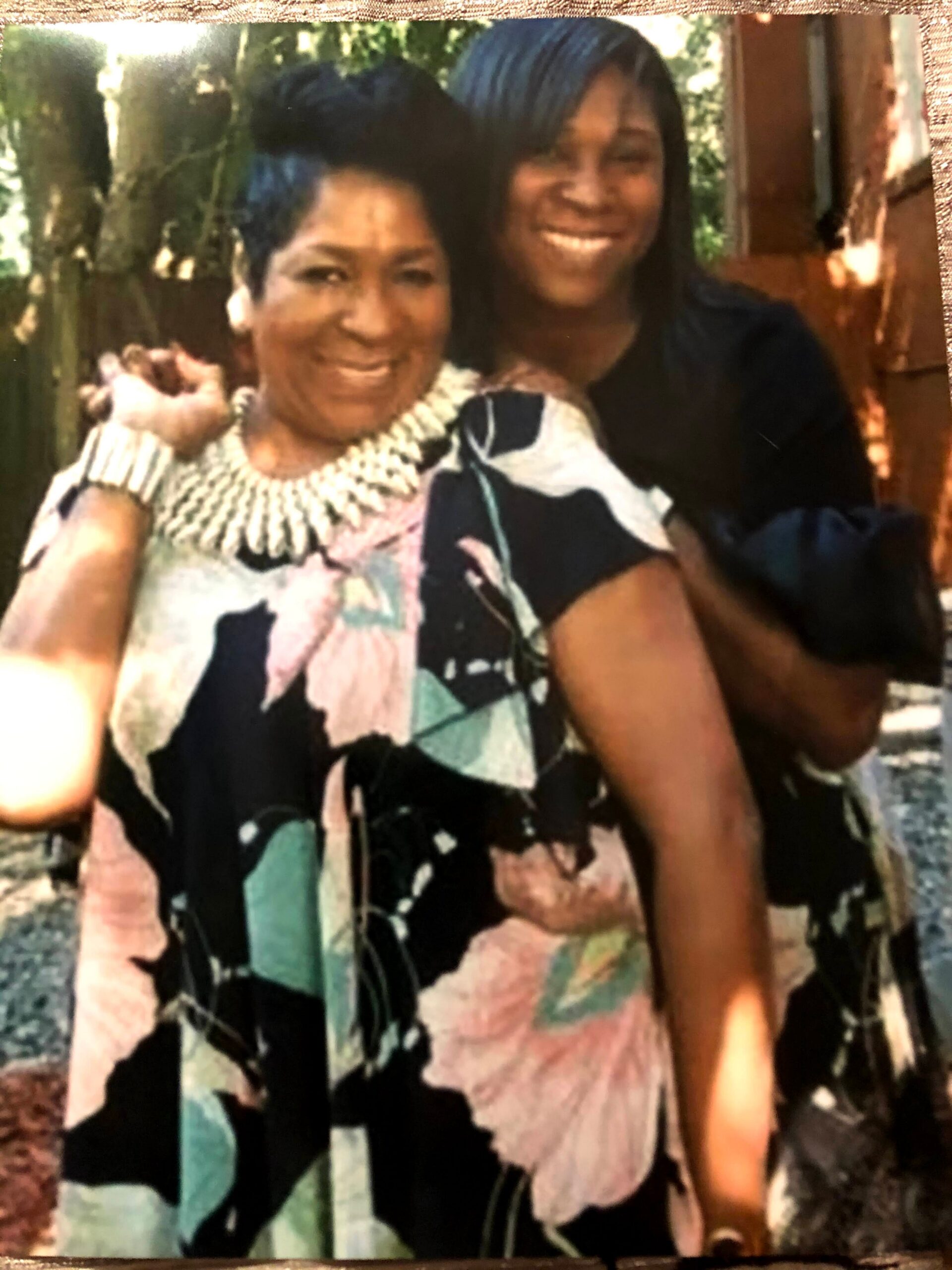 Two women posing together outdoors, smiling at the camera; older woman in a colorful patterned top stands in front, younger woman with blue hair stands behind her.