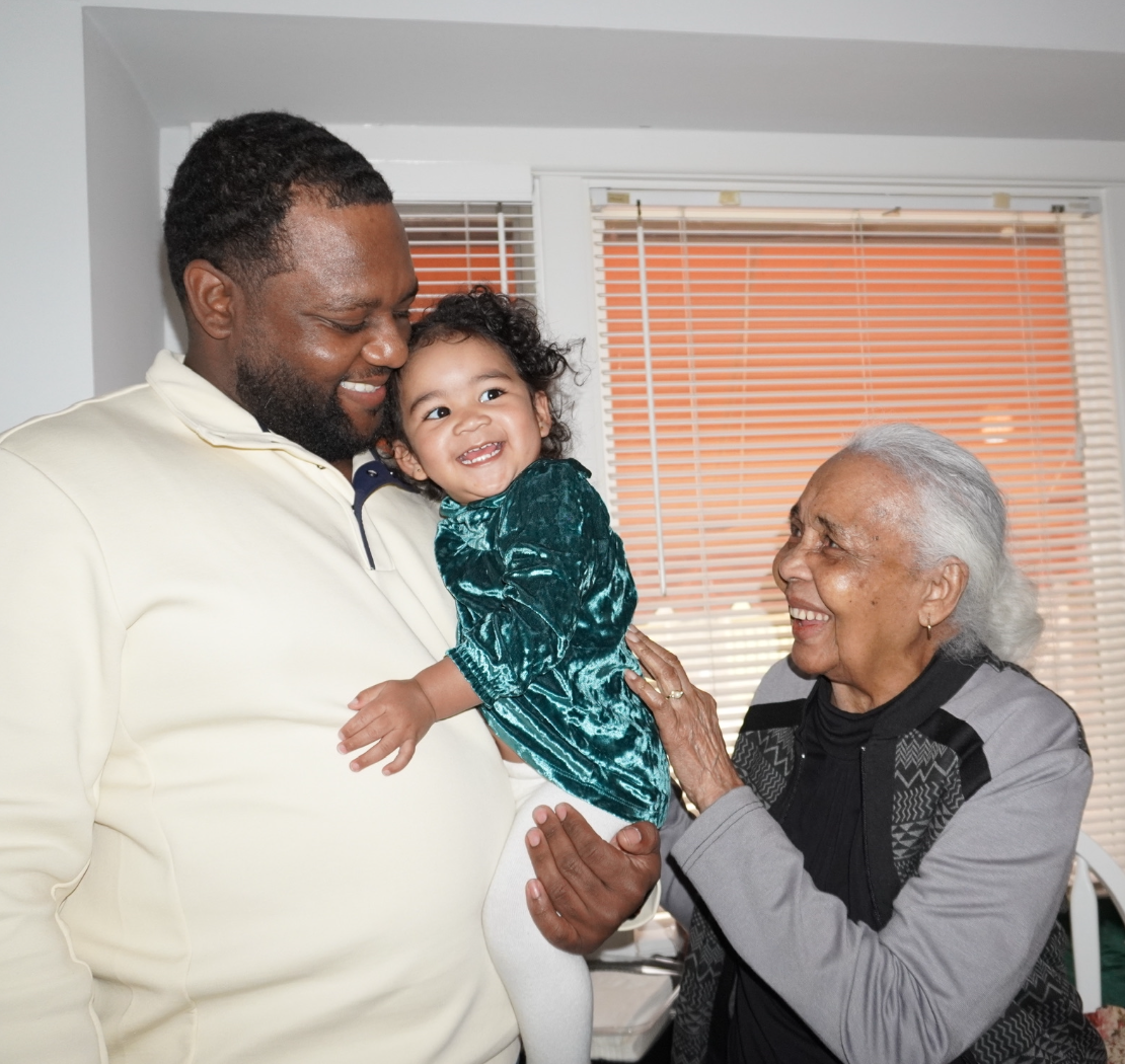 Smiling man holds a cheerful toddler as an elderly woman in a patterned cardigan smiles at them.