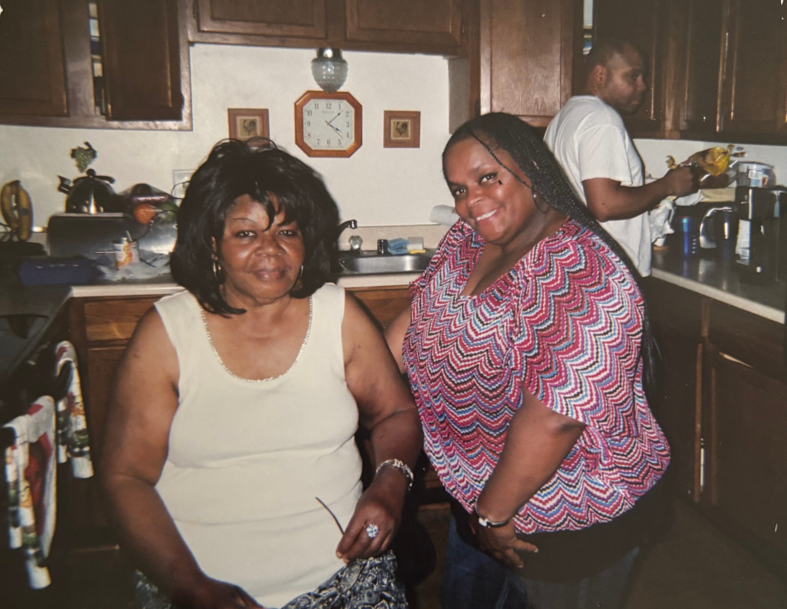 Two women posing in a kitchen, one in a light sleeveless dress and the other in a pink zigzag blouse, with a man in a white shirt at the counter in the background.