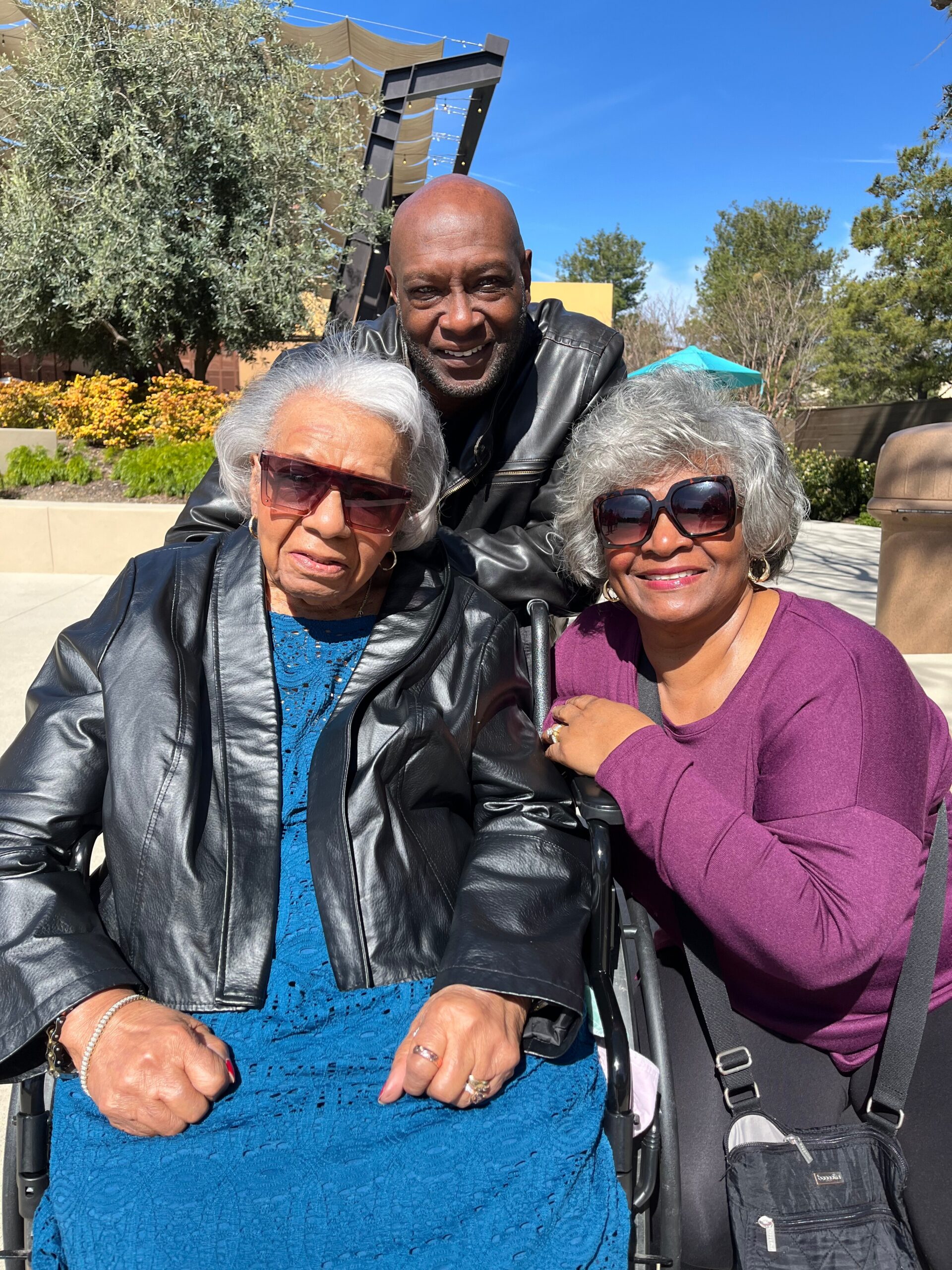 Three adults outdoors pose for a photo: two elderly women in wheelchairs wearing sunglasses, with a smiling man standing behind them on a sunny day.