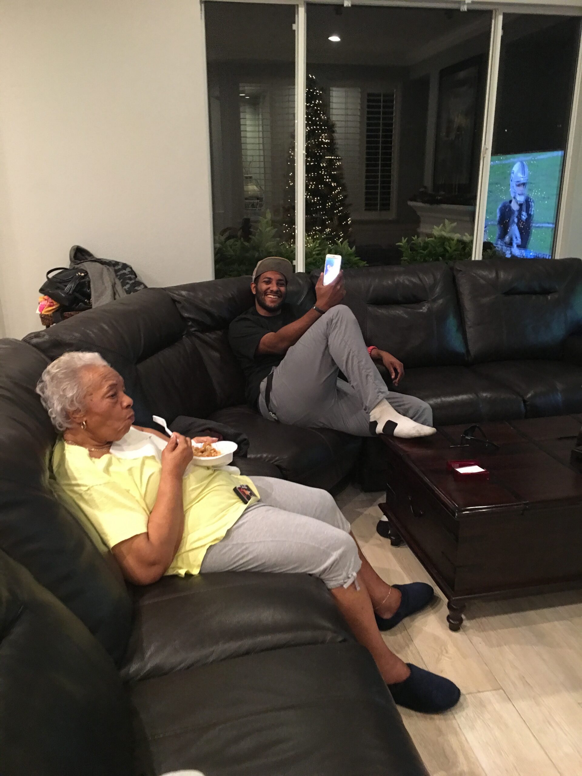 Man on a black leather sofa takes a selfie with his phone while an elderly woman beside him eats from a bowl in a living room.