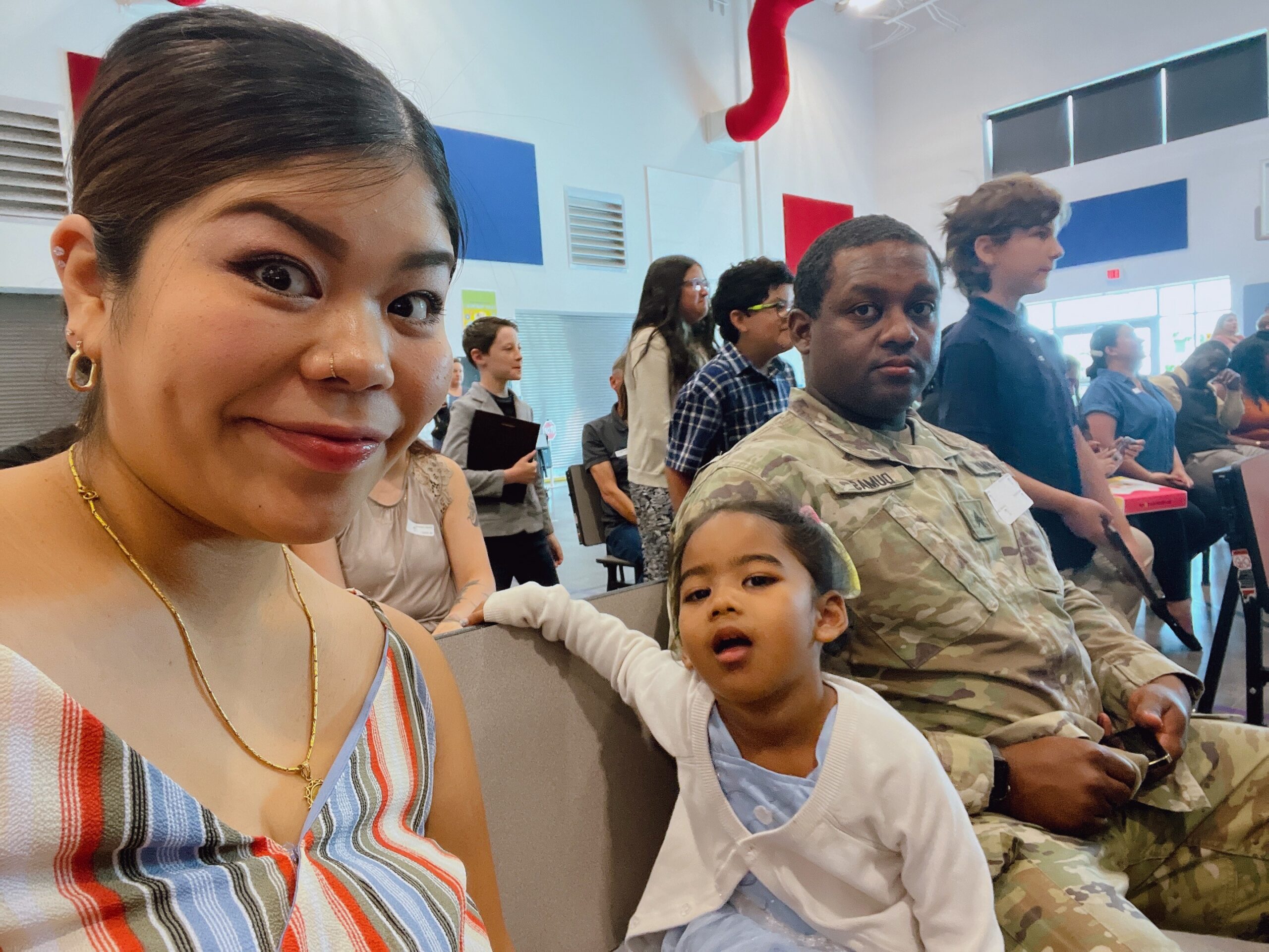 Smiling woman in a striped dress taking a selfie in a crowded waiting area; a child in a white coat sits beside her while others wait in the background.
