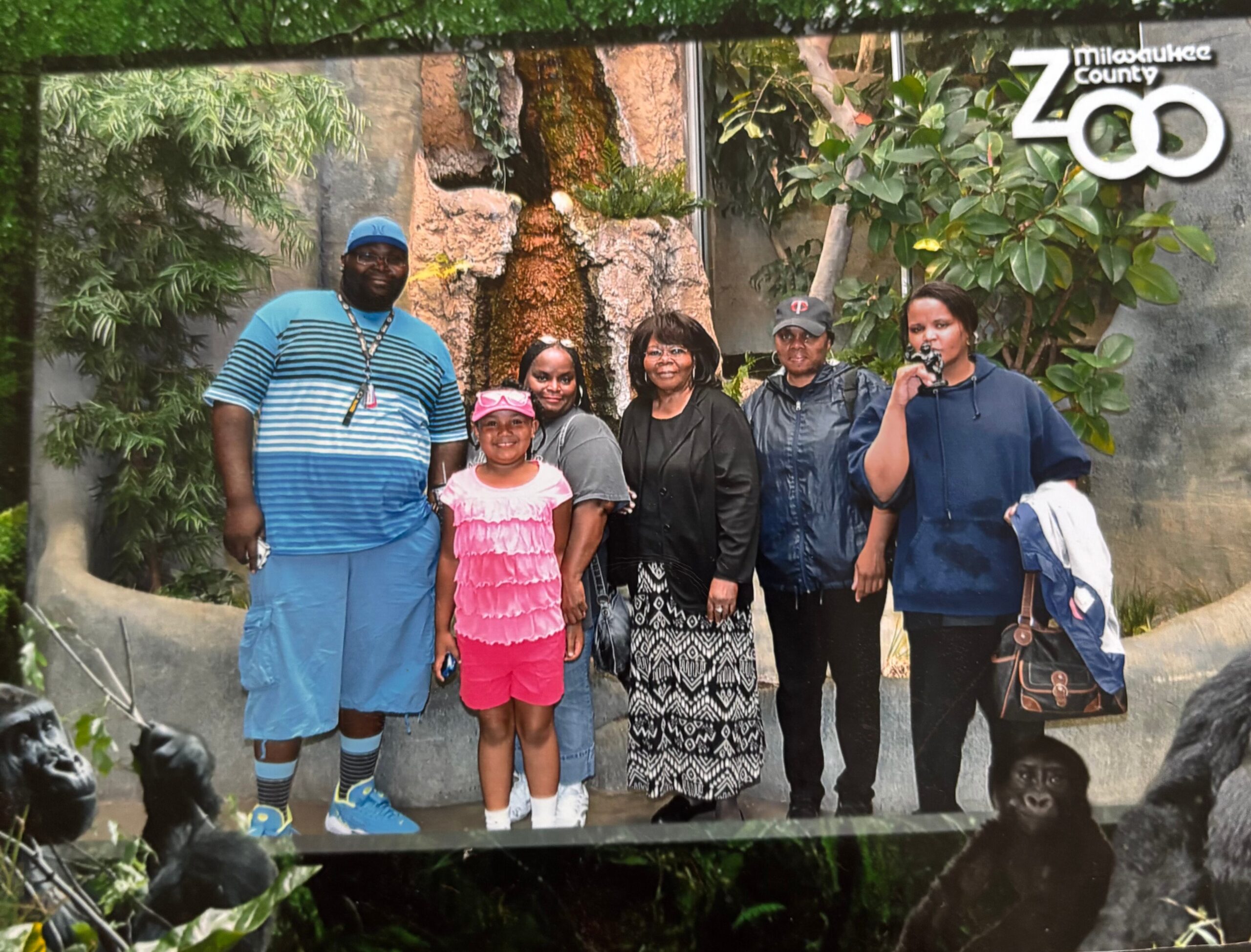 Group of six people posing outdoors at Milwaukee County Zoo, with a carved tree sculpture in the background.