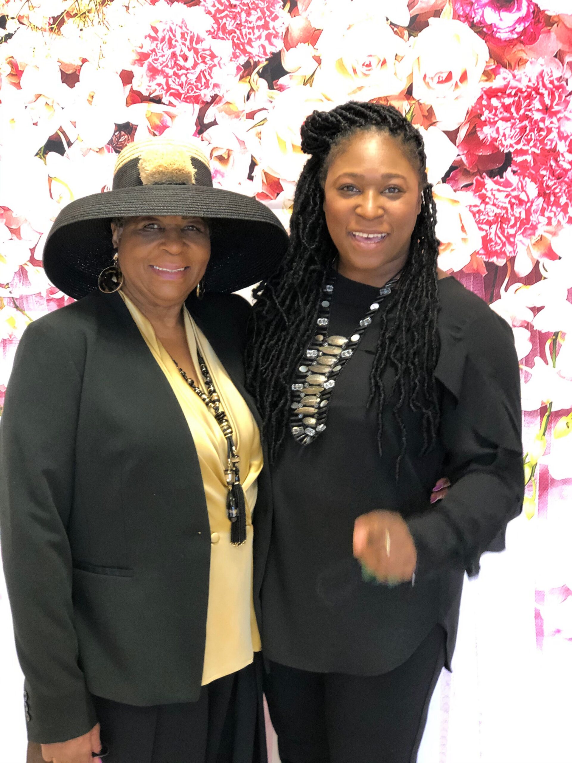 Two women posing together in front of a pink floral backdrop; older woman on the left wears a black hat and blazer with a pale blouse, younger woman on the right wears black with a beaded necklace.