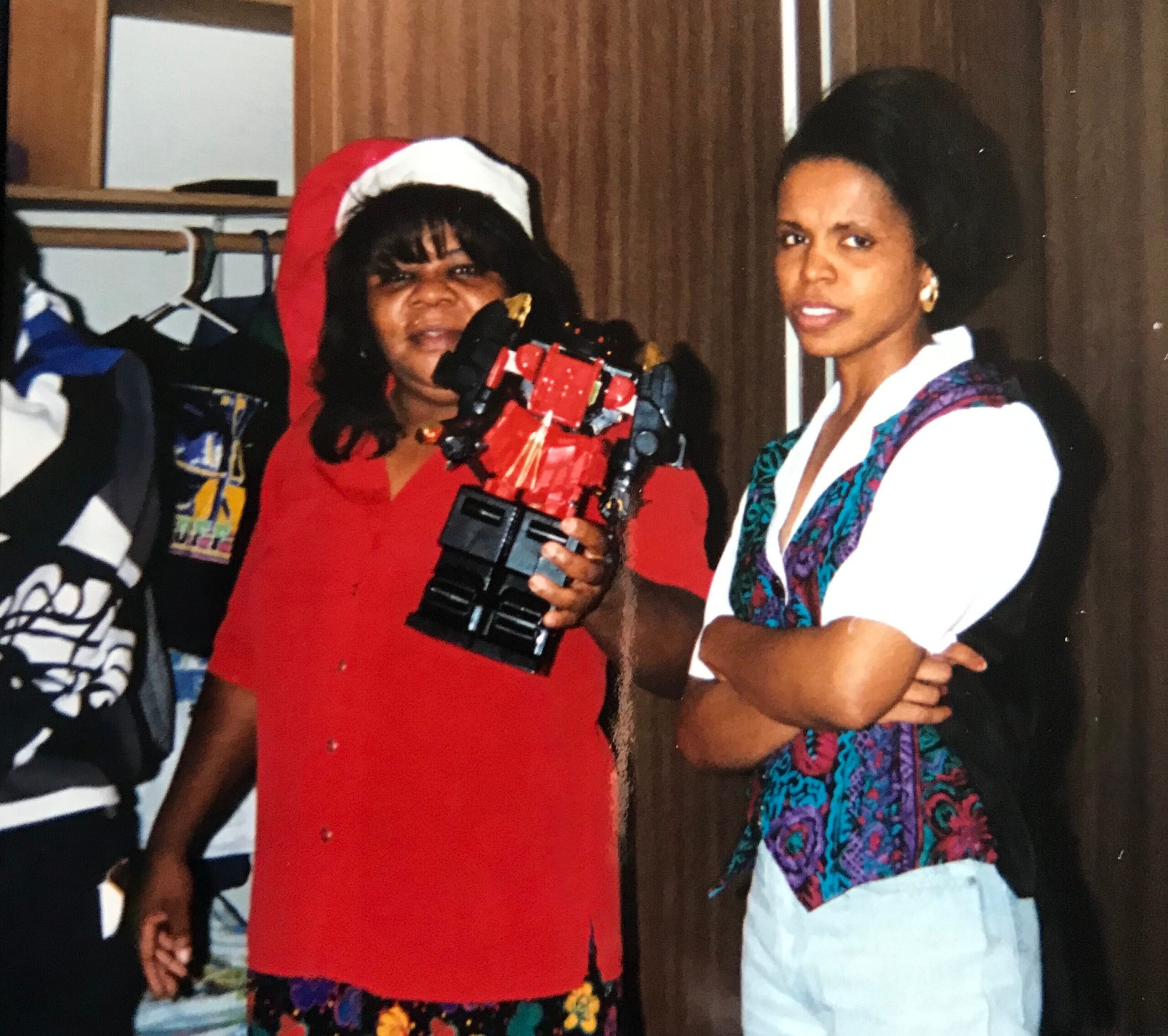 Two women posing for a photo indoors; one in a red dress with a Santa hat holding a video camera setup, the other in a white top with a colorful scarf.