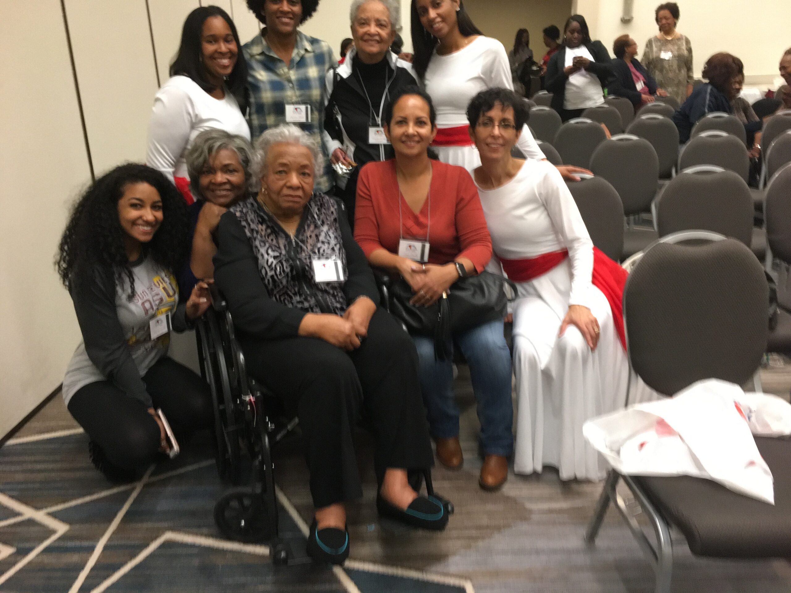 Group of diverse women posing together in a conference room; some seated, some standing behind, all wearing name badges.