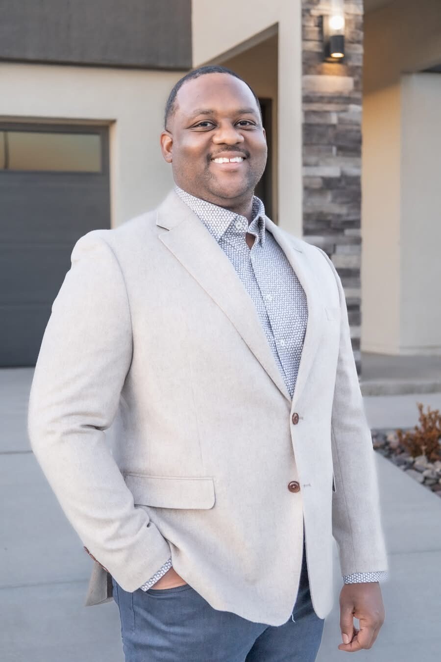 Smiling man in a light beige blazer and patterned shirt stands outdoors in front of a modern home with a garage door visible behind him.
