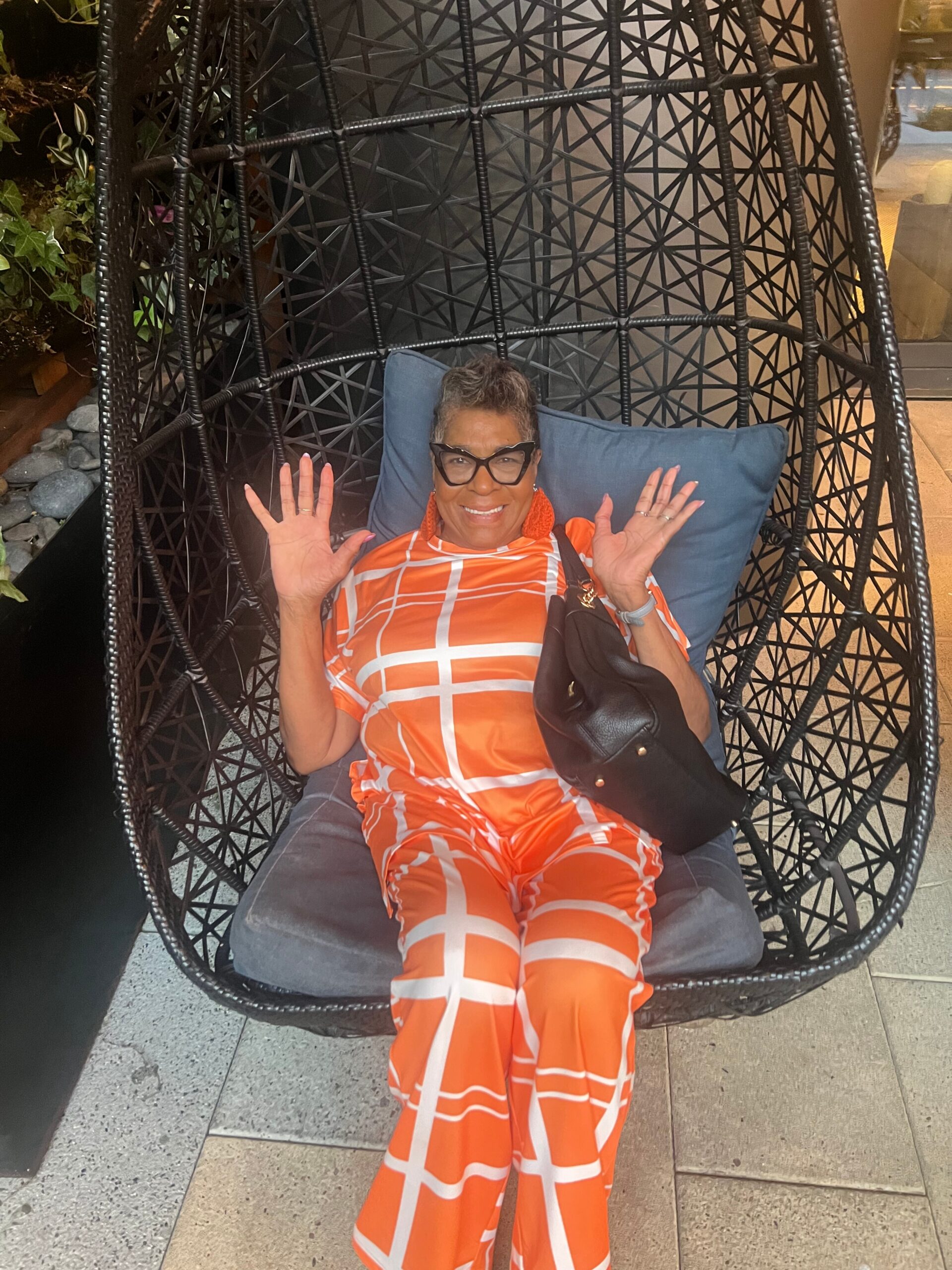 Smiling woman with short gray hair and glasses sits in a black wicker hanging chair with a blue cushion, wearing an orange geometric outfit and raising both hands in a greeting gesture.