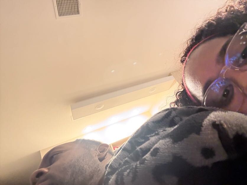 Close-up selfie of two people close together in a room, one wearing clear glasses and a red headband, the other partially visible under a patterned blanket with a pale background ceiling light behind them.
