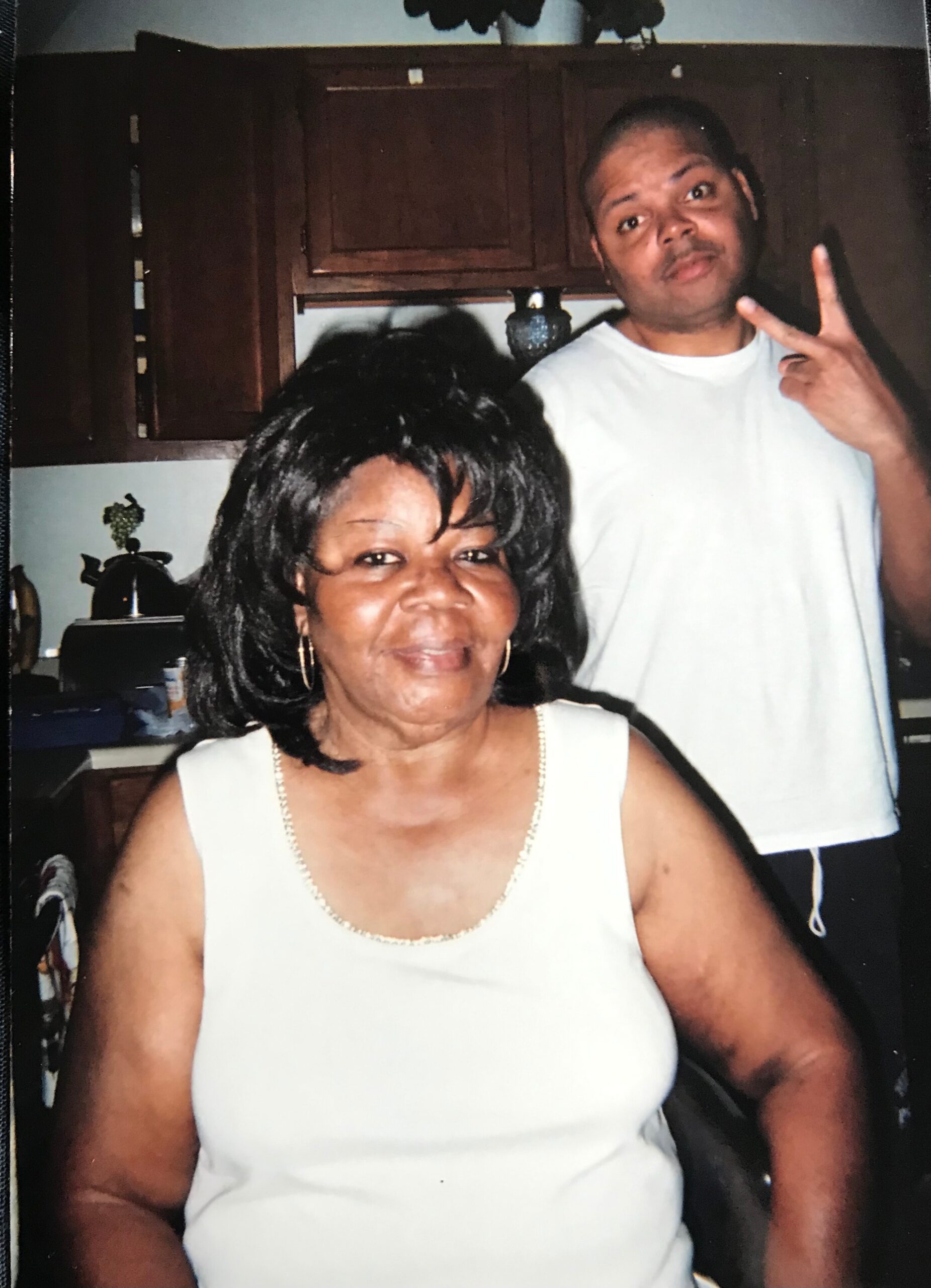 Smiling woman in a white sleeveless top in a kitchen, with a man in a white shirt behind her making a peace sign, posing for a photo.