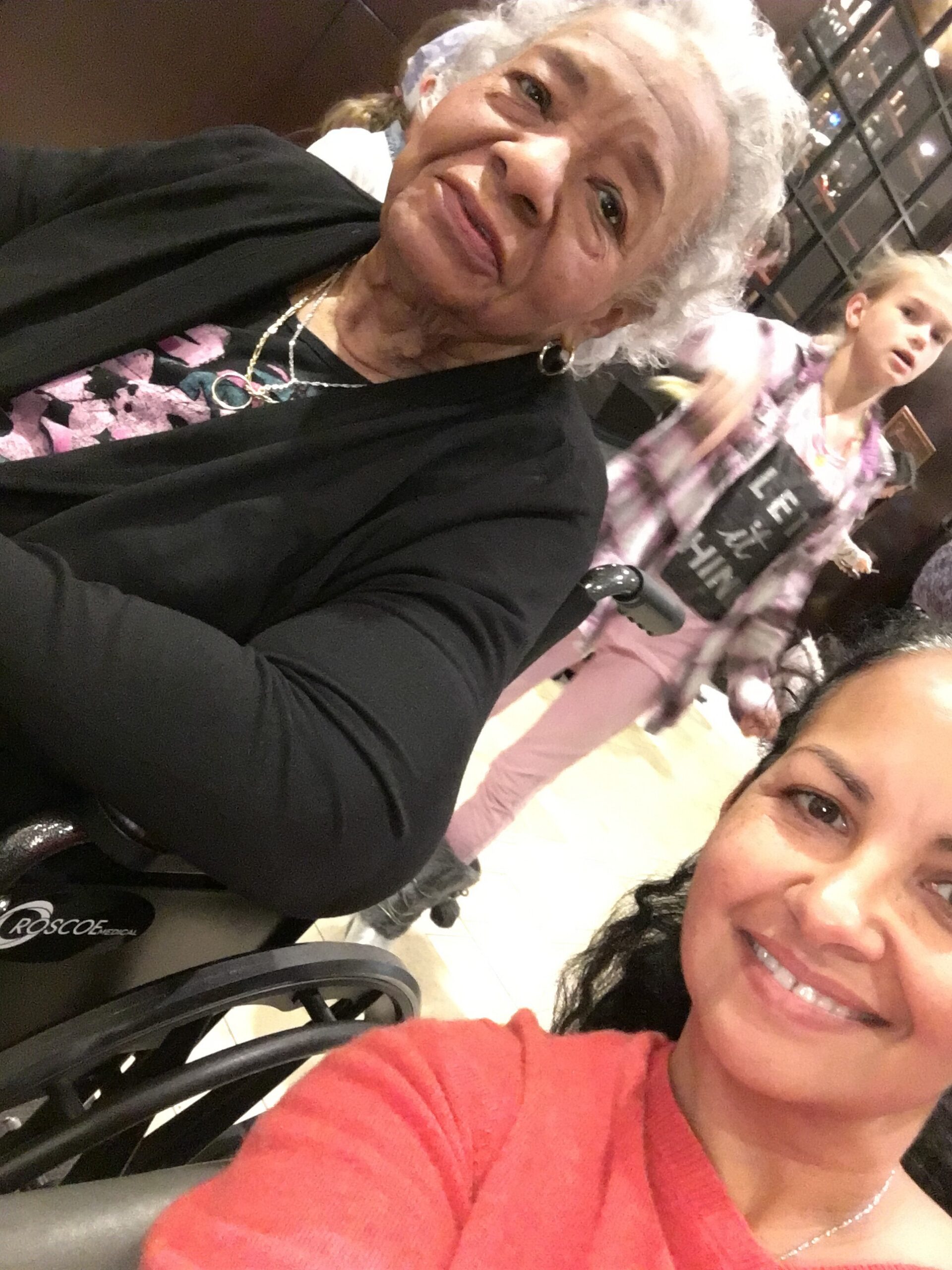 Two women posing for a selfie in a store; foreground shows a woman in a pink sweater smiling, an older woman with white hair in a black jacket beside her, and a wheelchair visible in the foreground.