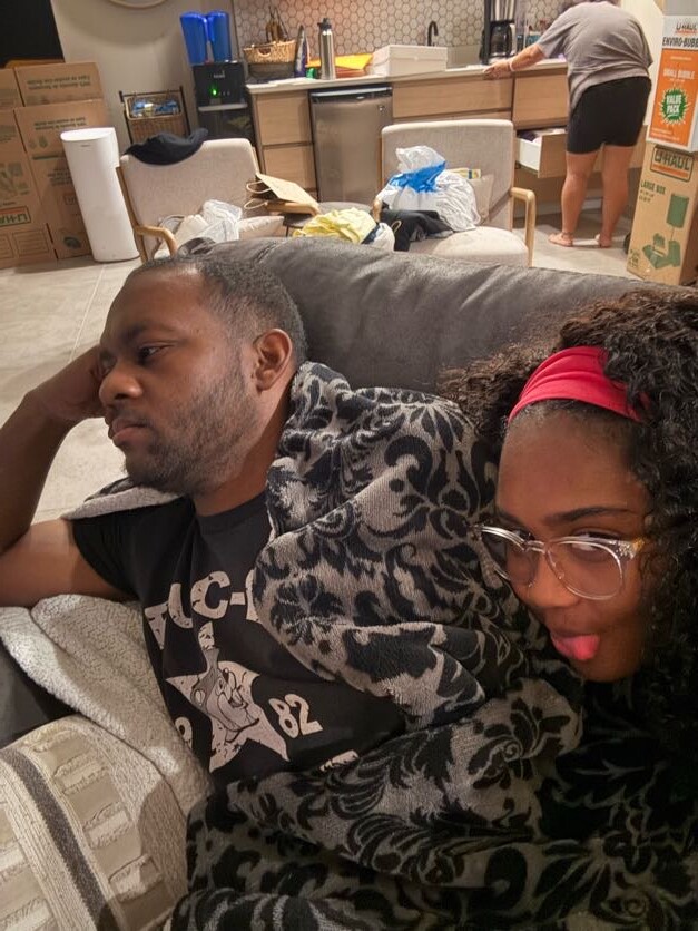 Man and woman cuddled on a gray couch, covered with a black-and-gray patterned blanket; the woman wears a red headband and glasses, sticking out her tongue. In the background, a cluttered kitchen with boxes and a person standing at the counter.