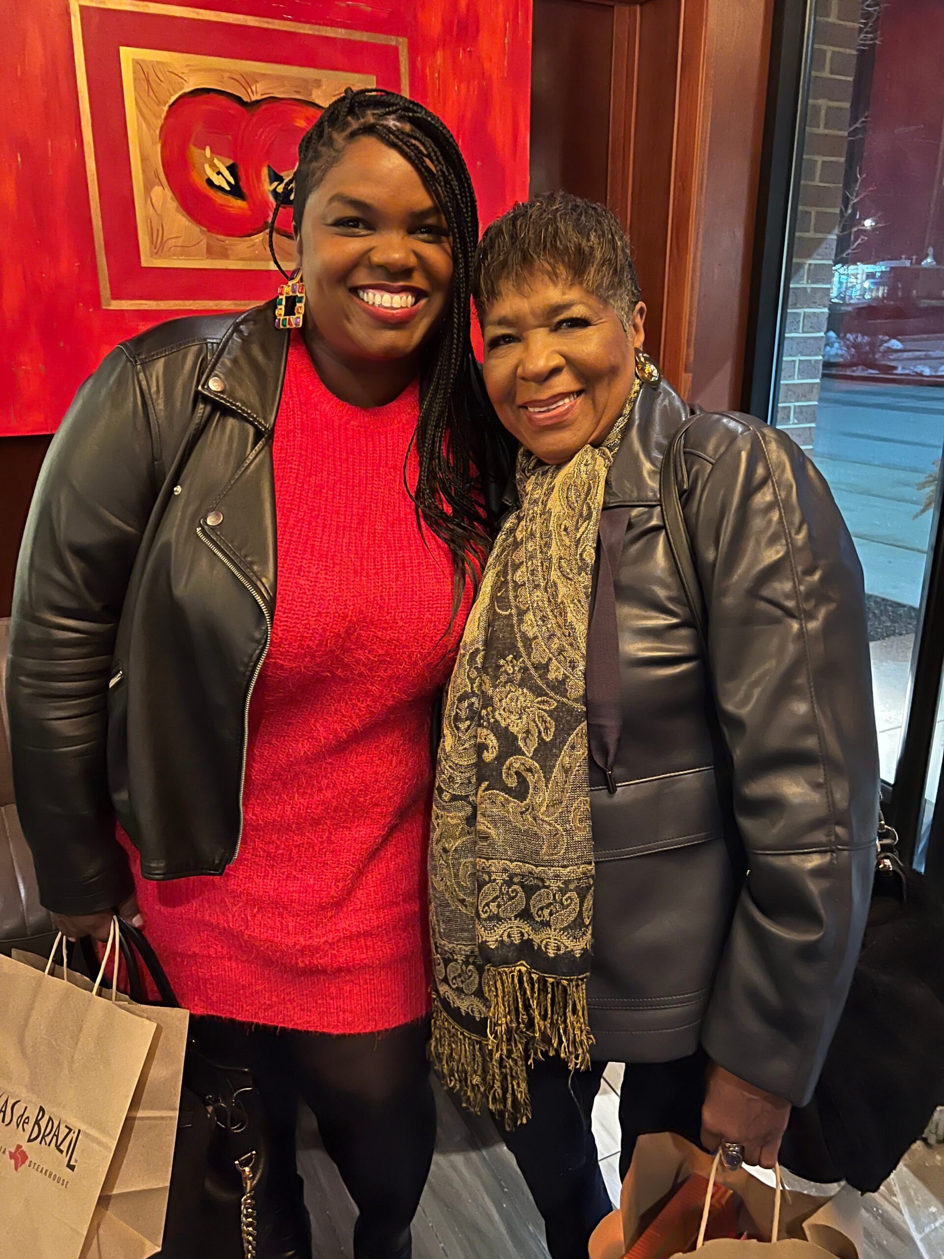 Two women smiling for the camera indoors, wearing leather jackets. One in a red sweater with braided hair, the other in a patterned scarf, both holding shopping bags.