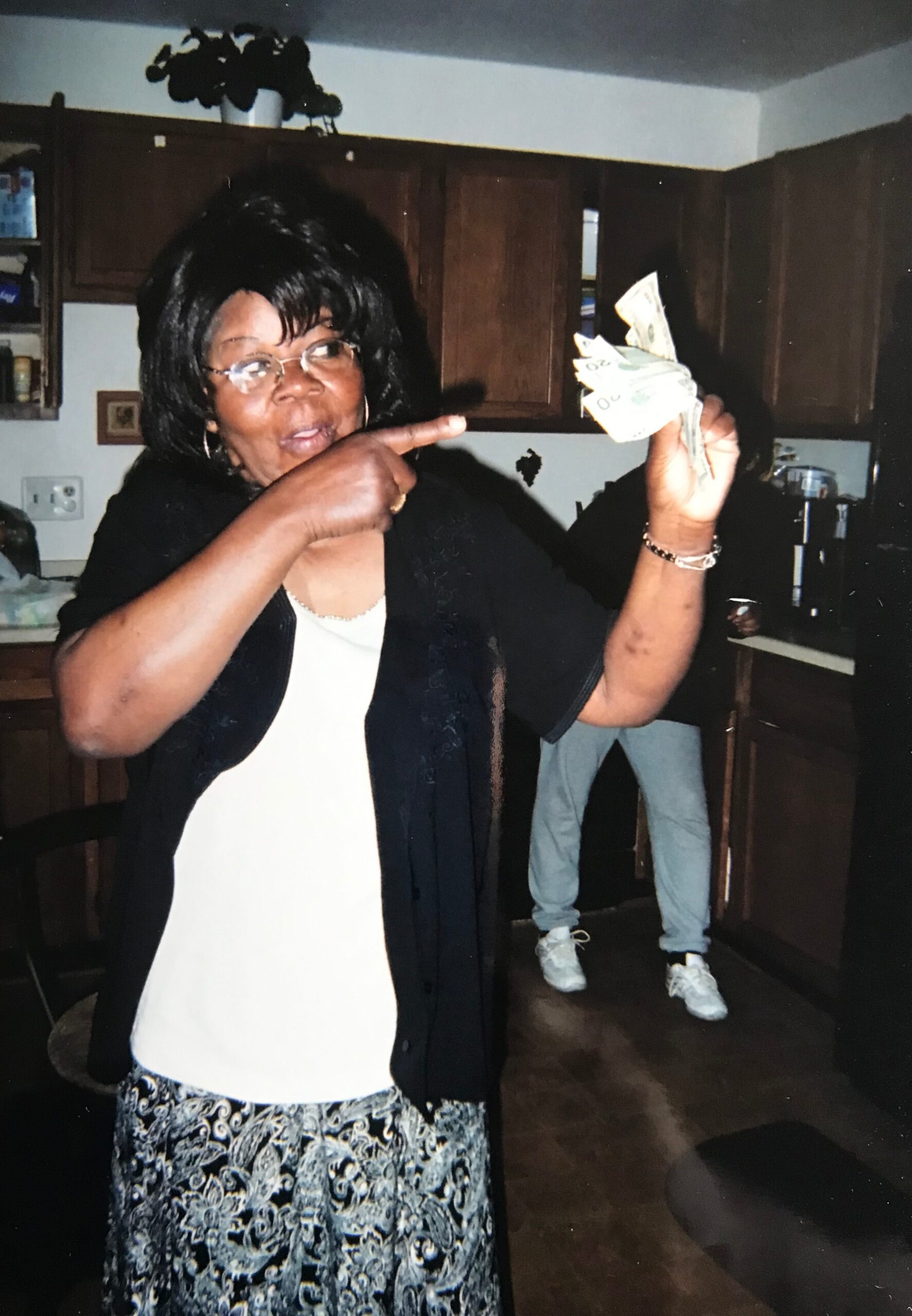 African American woman with glasses in a kitchen points at a fan of folded cash held in her right hand.