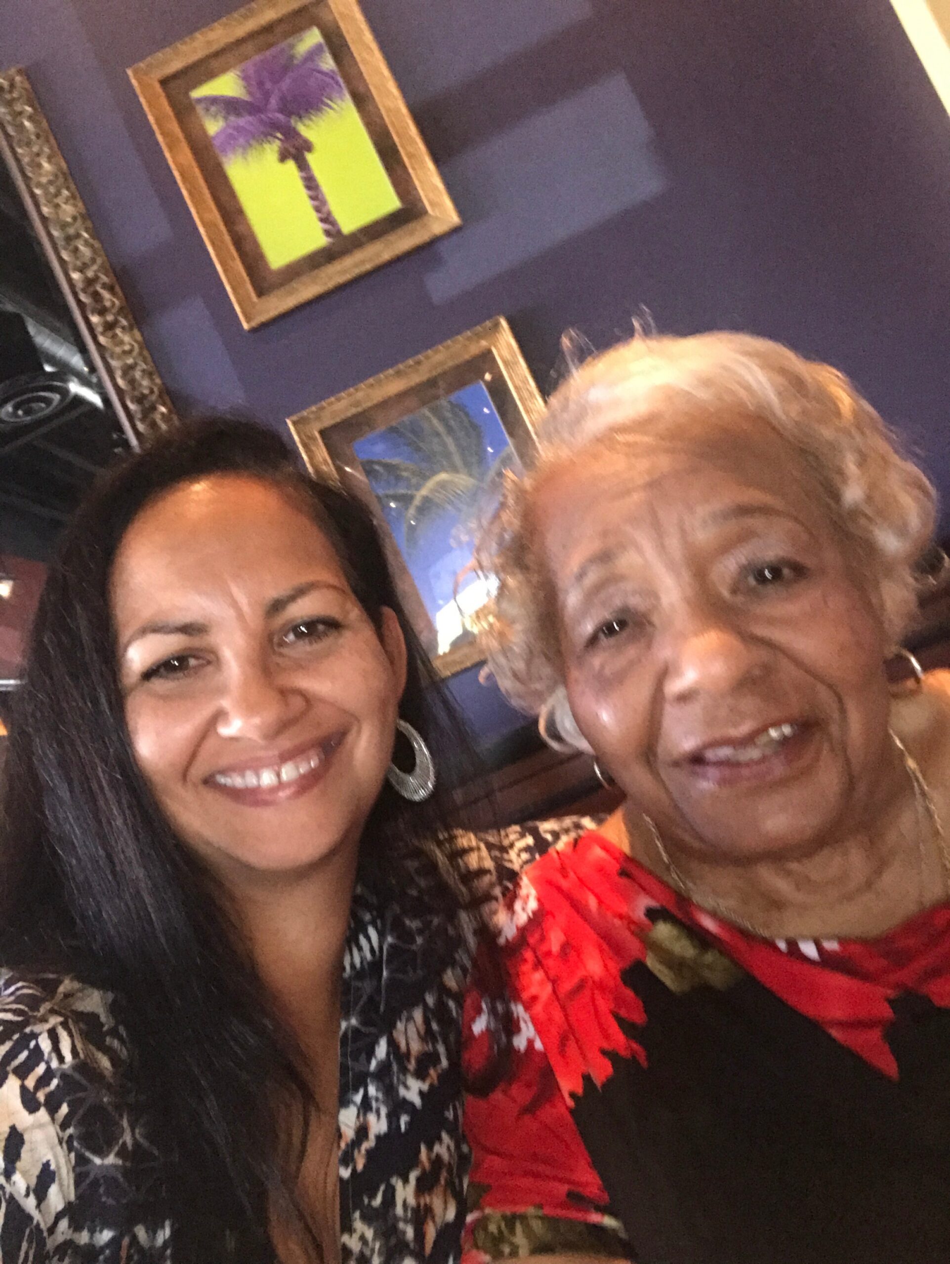 Two women smiling for a selfie against a purple wall with framed palm-tree pictures in the background.