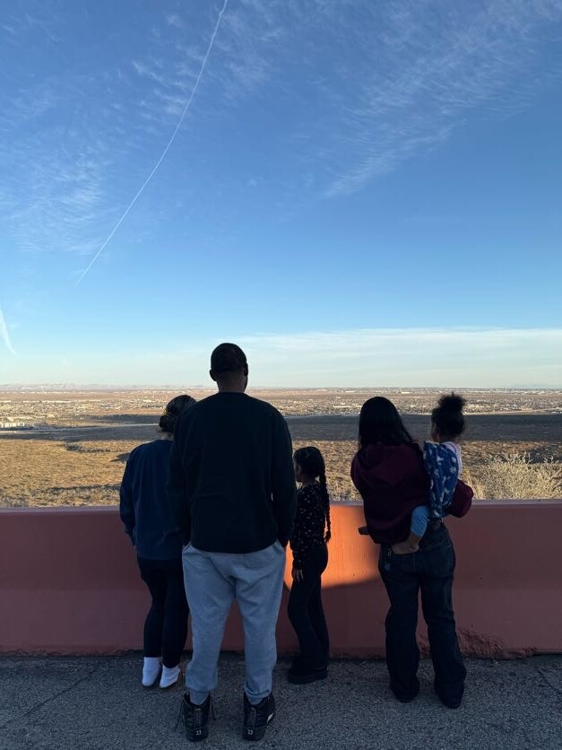 Family of five leaning on a pink railing, looking out over a desert valley toward a distant city under a clear blue sky with a contrail.