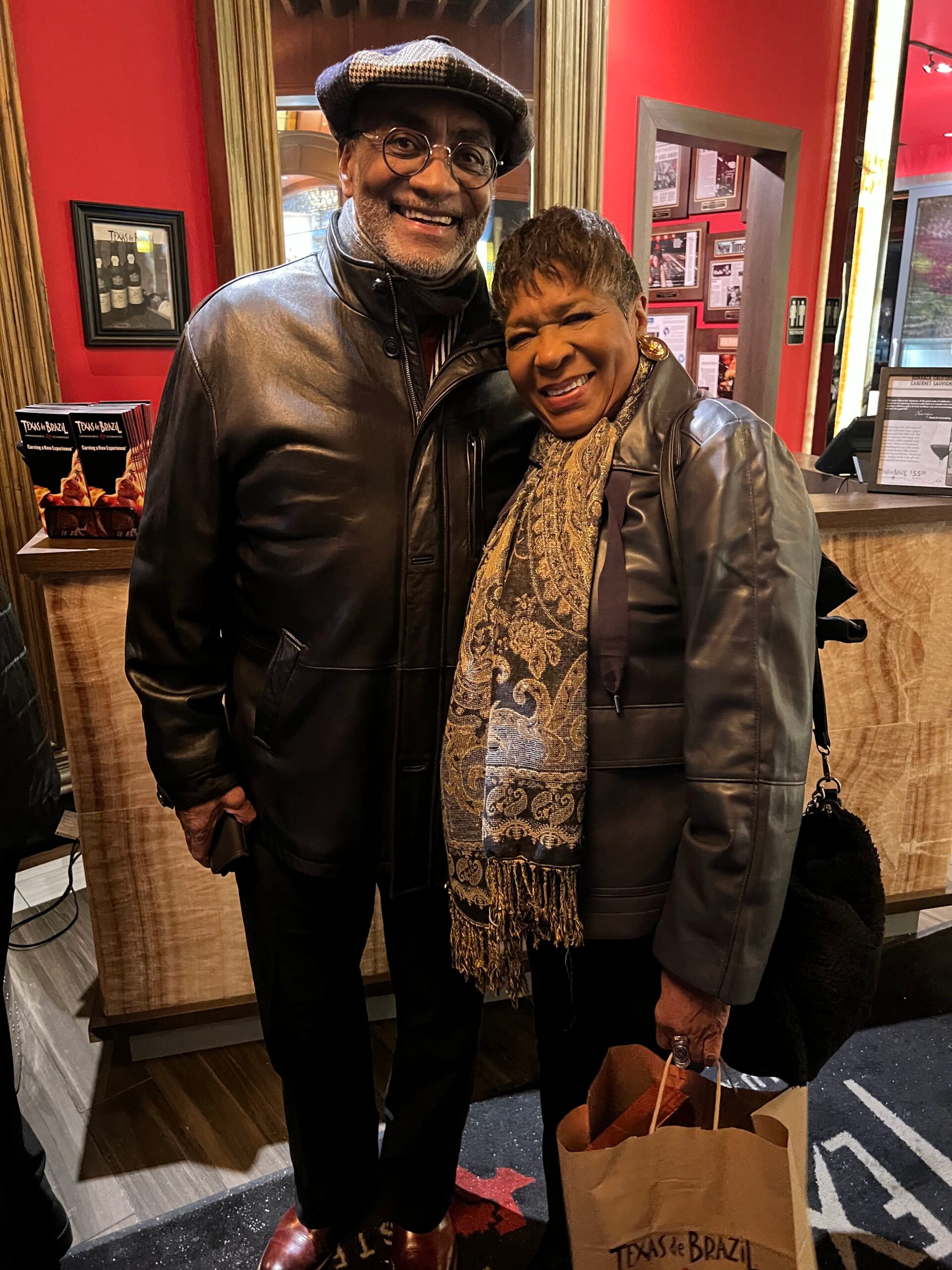 Two smiling adults pose together indoors in a restaurant lobby; man in a leather jacket and cap, woman in a leather jacket with a patterned scarf, standing close together with a Texas de Brazil bag nearby.