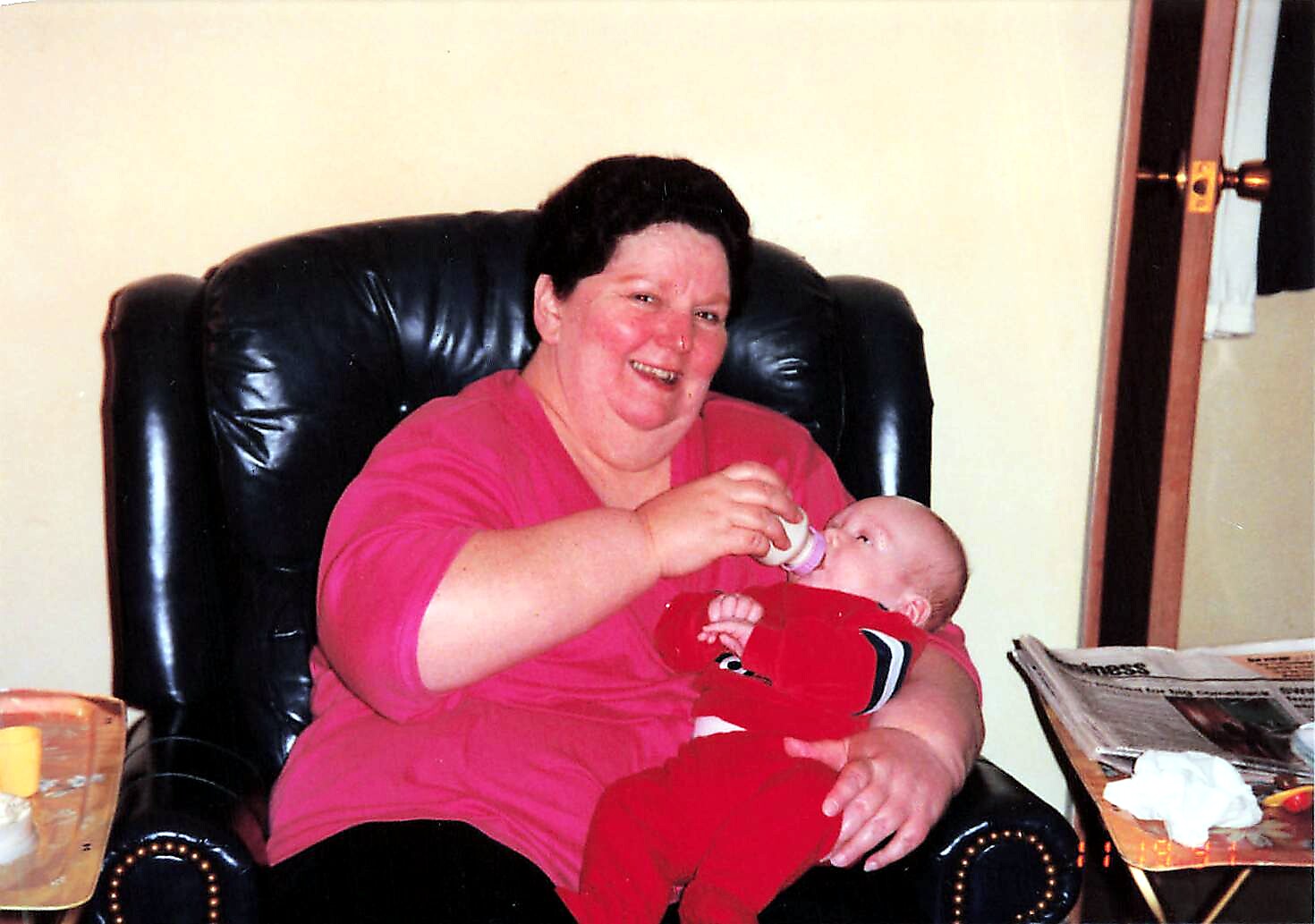 Woman in a pink top sitting in a black leather chair feeds a baby with a bottle in a living room setting.
