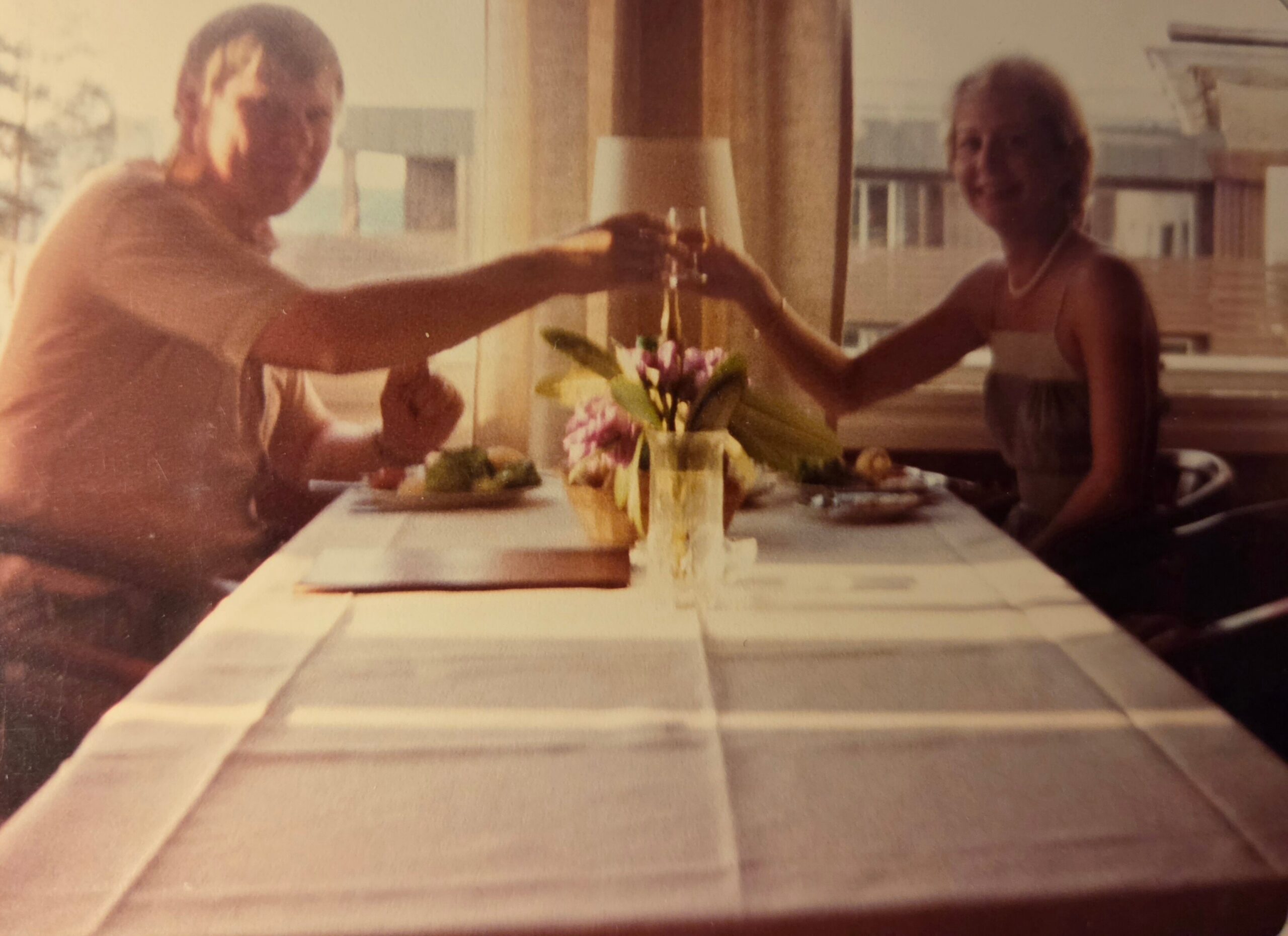 Two people toasting with glasses at a dining table with a flower centerpiece.