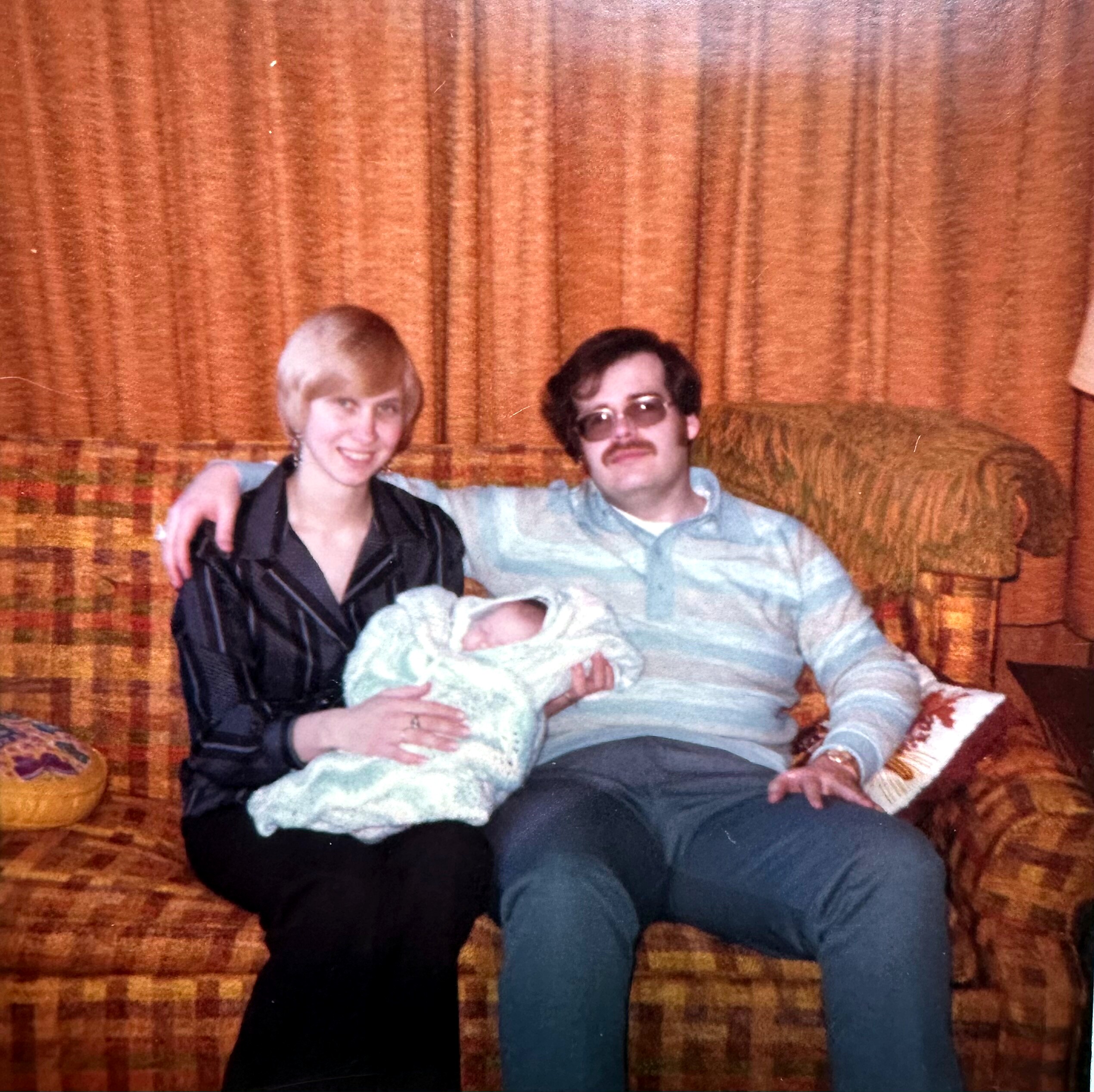 Two adults sit close on an orange plaid sofa; a baby wrapped in a light blanket rests on the woman's lap.