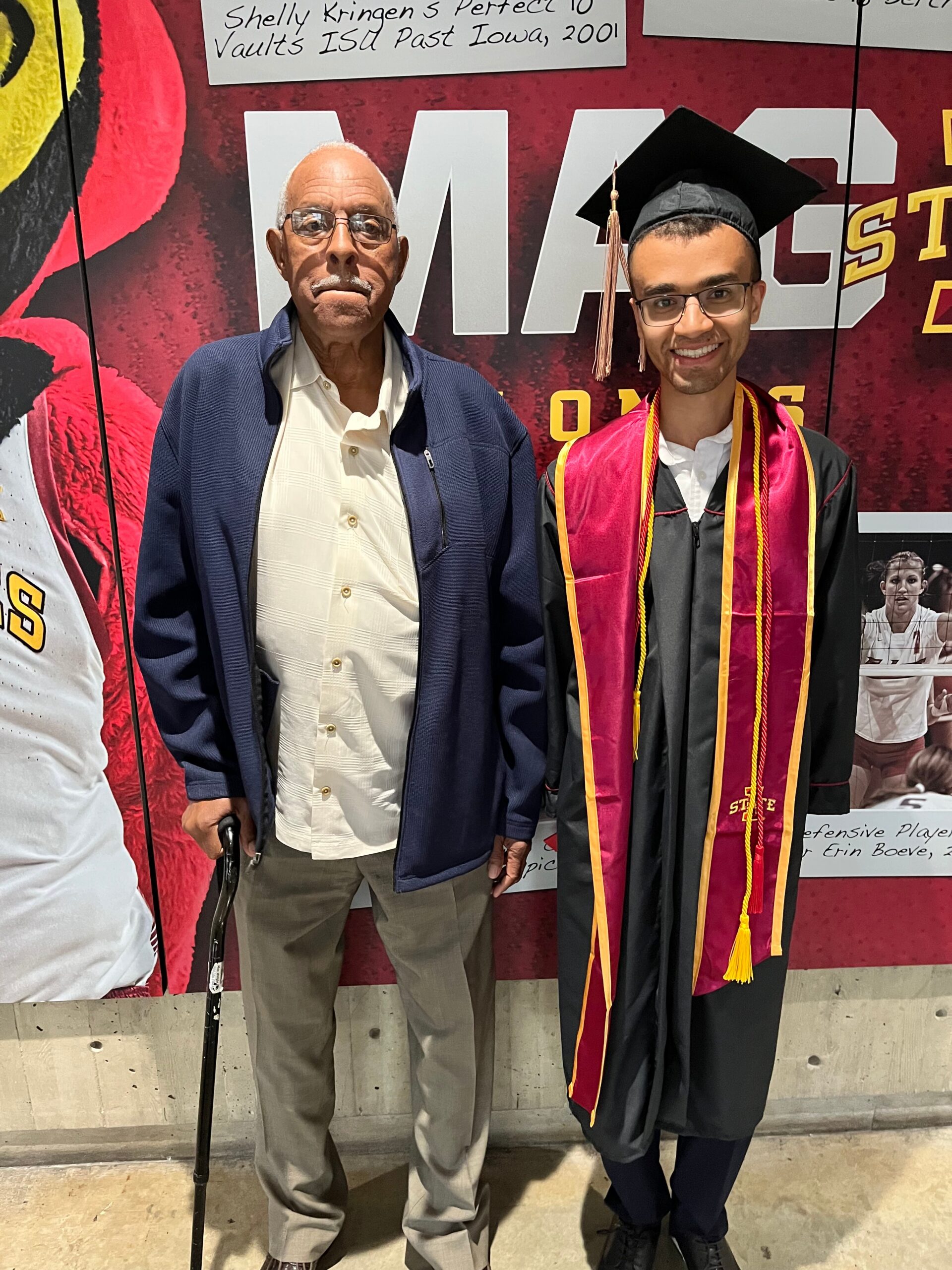 Two men posing indoors in front of a red backdrop: an older man in a blue jacket and light shirt with a cane, next to a smiling graduate in a black cap and gown with maroon and gold stole.
