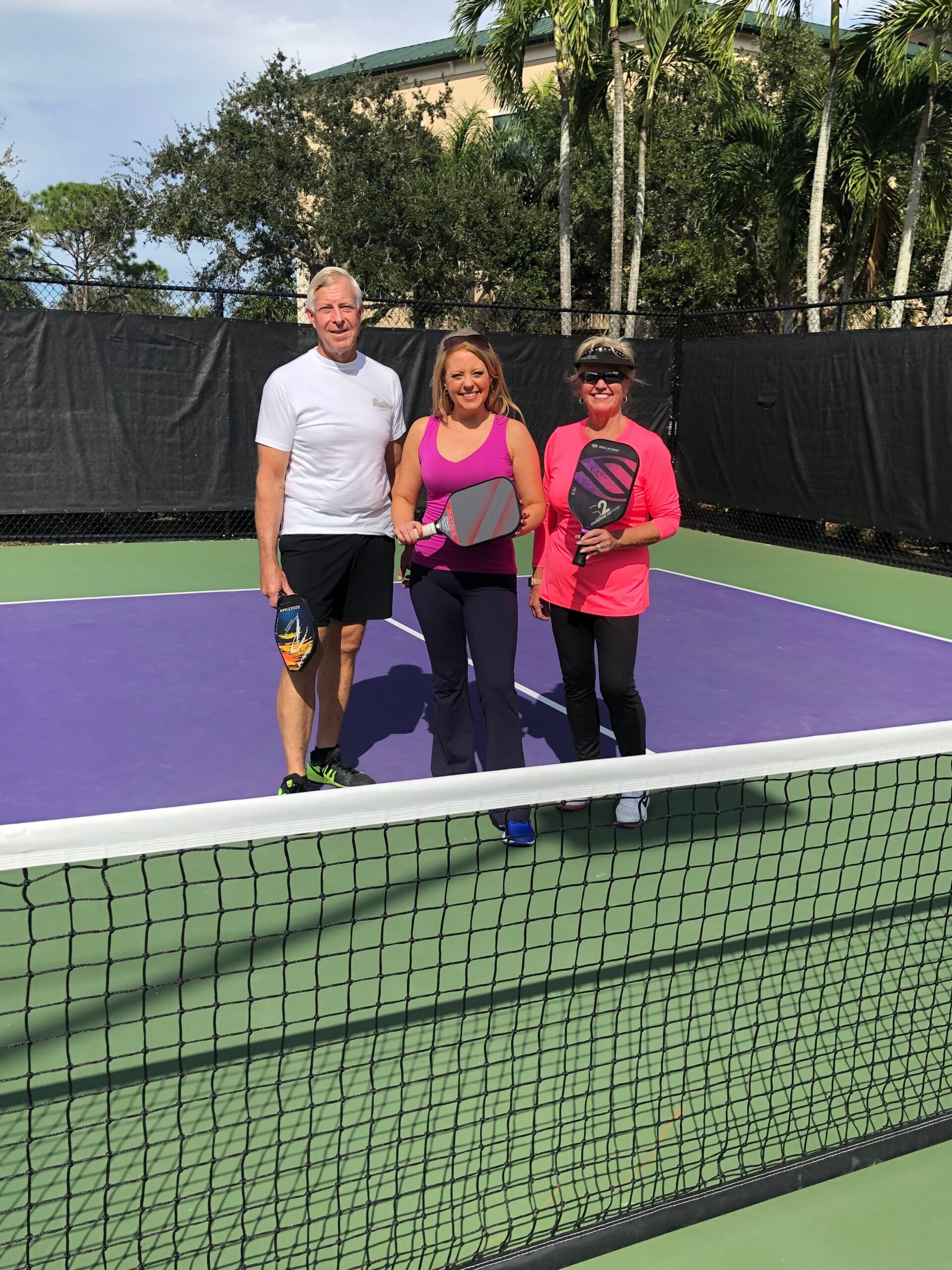 Three adults posing with paddles on a purple tennis/paddle court, standing side by side in front of the net.