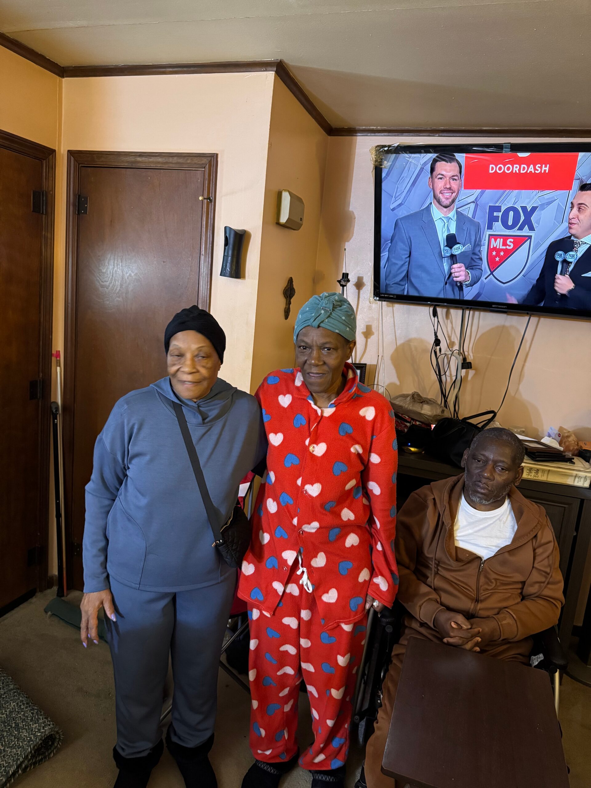 Three adults in a living room: two standing women in colorful pajamas and a man in a brown jacket seated, watching TV together with a Doordash/Fox MLS broadcast on the screen above.