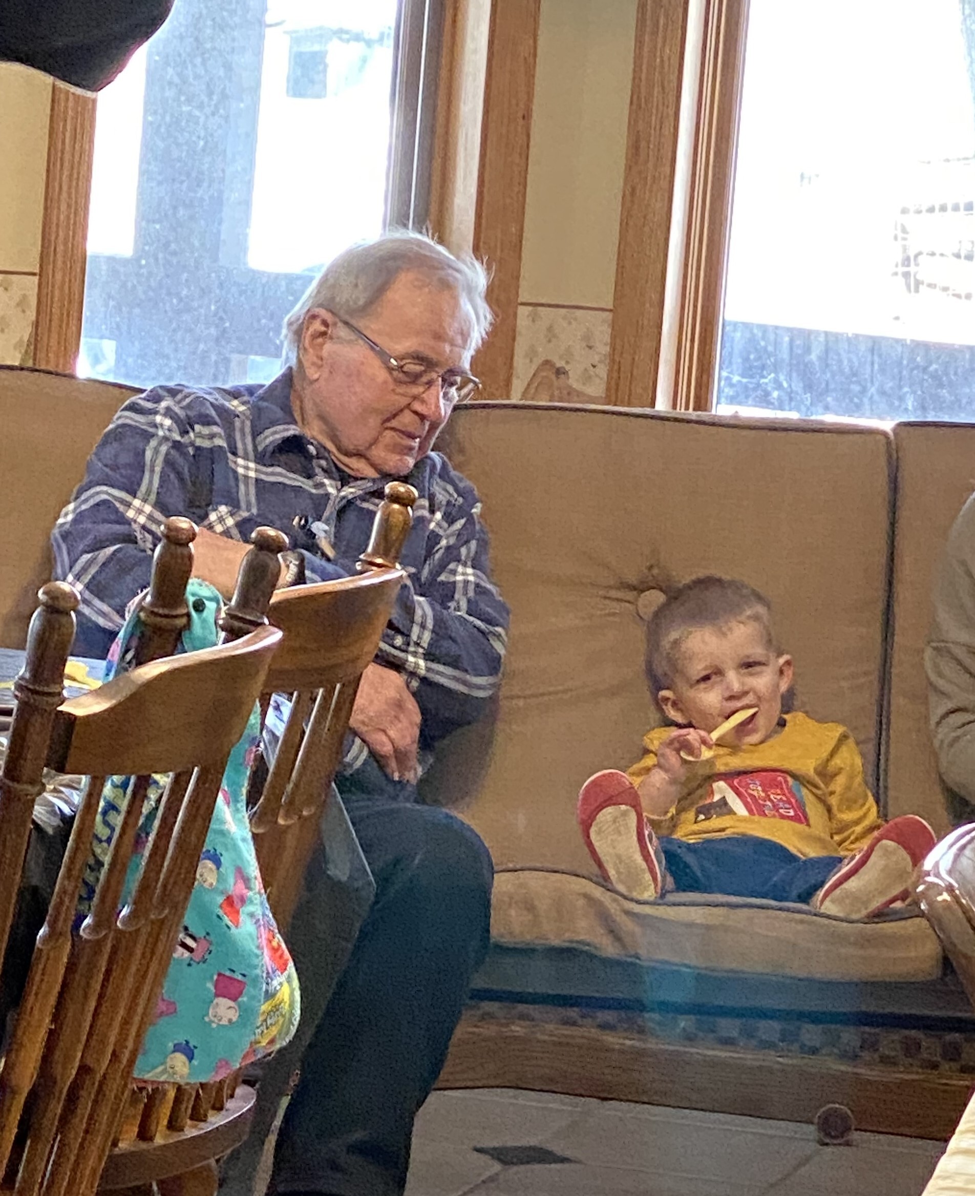 Older man with glasses and gray hair sits on a sofa, looking down at something in his lap while a young child in a yellow outfit sits beside him indoors near a window.