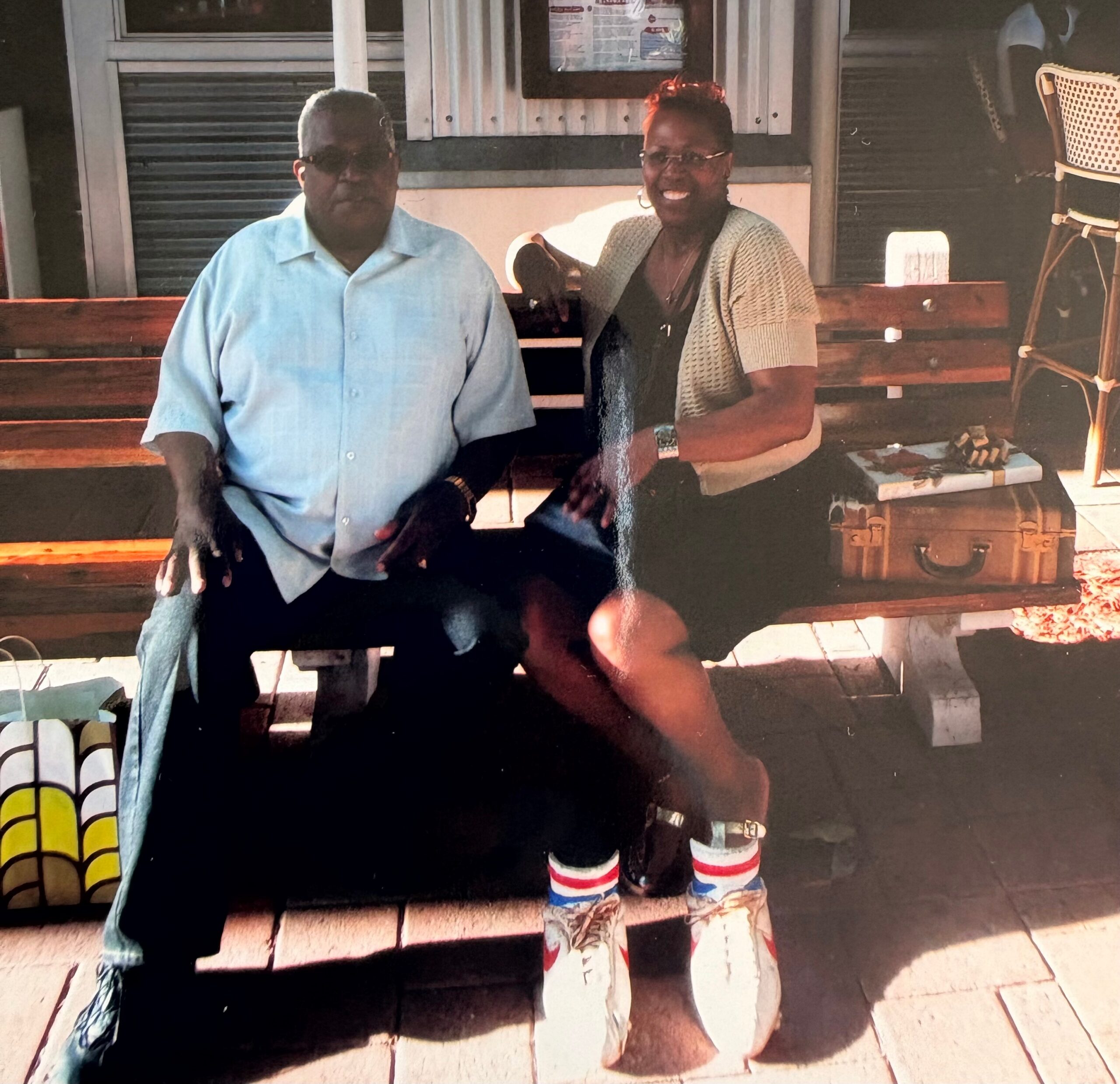 Two adults seated on wooden steps outdoors; man in a light blue shirt on the left and woman in a cardigan with glasses on the right, smiling at the camera.