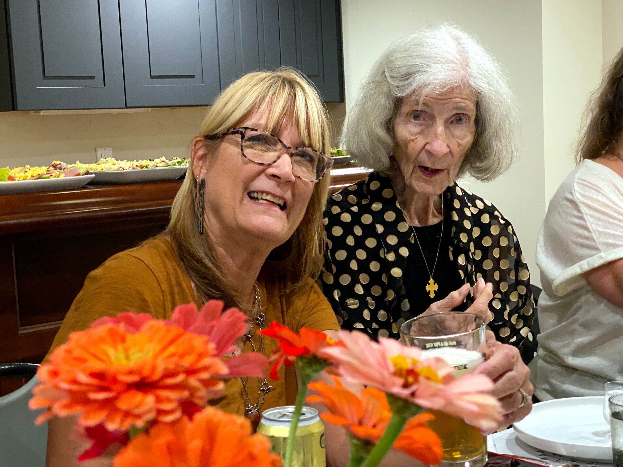 Two smiling women sit at a table with bright orange flowers in the foreground, in a kitchen-dining area behind them.