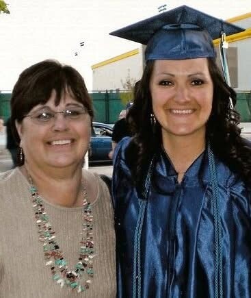 Graduate in blue cap and gown posing with a smiling woman in a beige sweater outdoors.