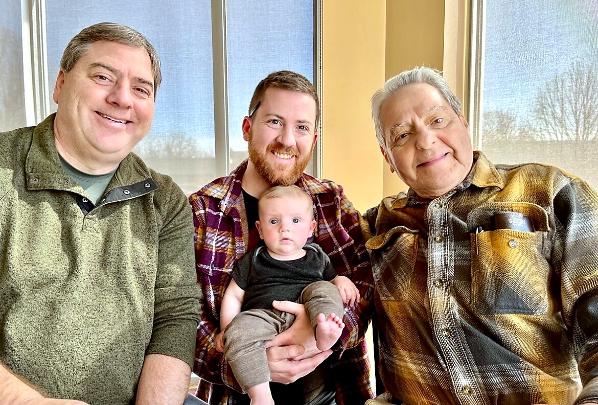 Three men smiling for a photo with a baby held by the man in the center, indoors by large windows.