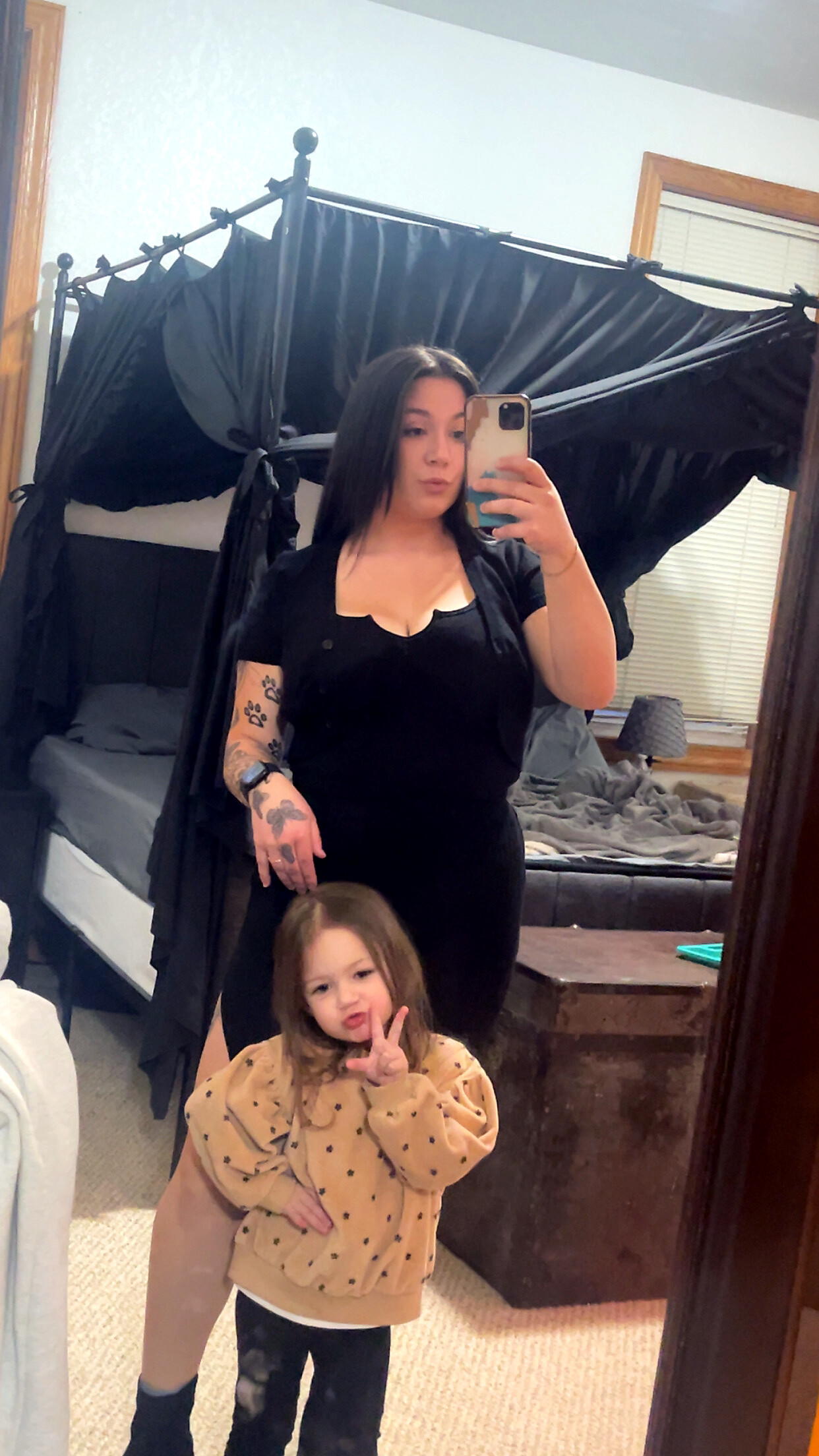 Woman in black outfit taking a mirror selfie in a bedroom; a young girl in a tan sweater stands in front, making a peace sign.