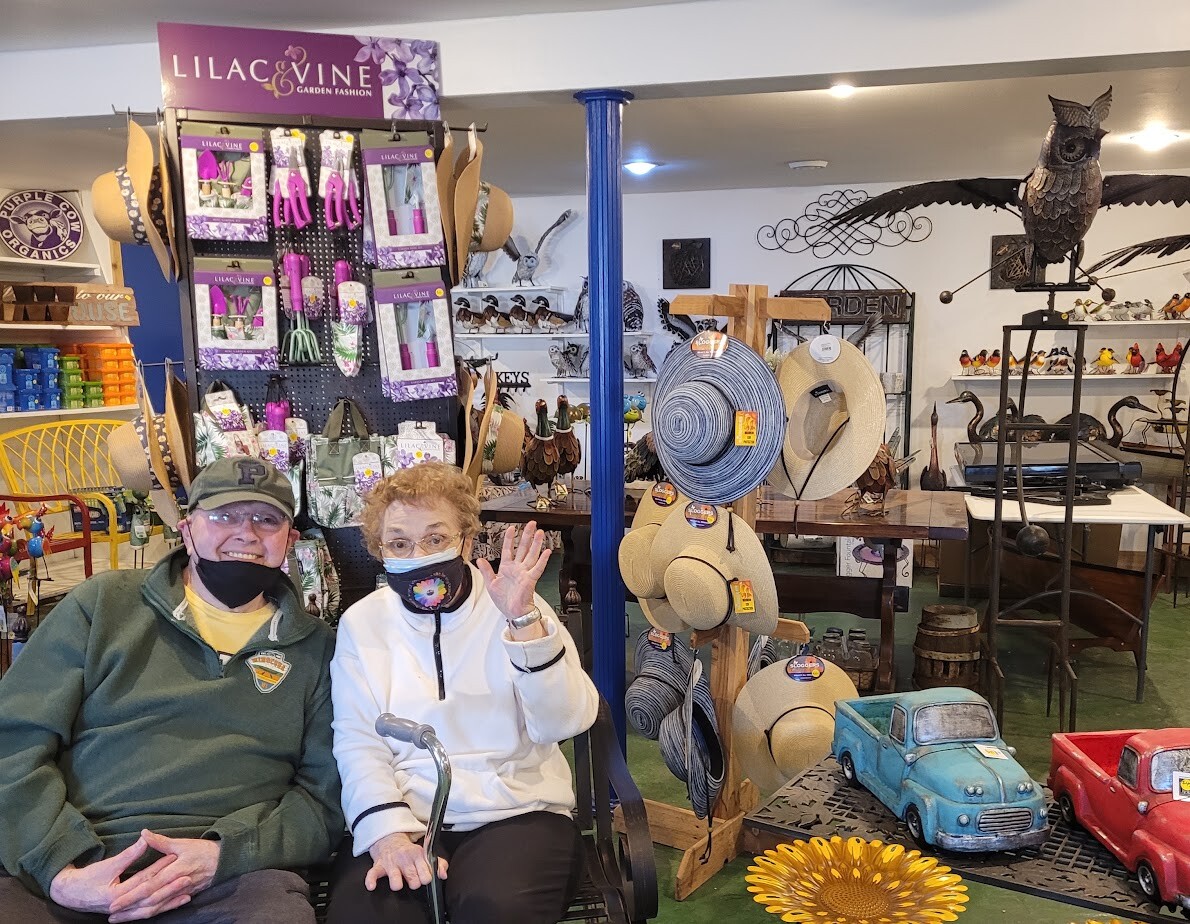 Two seniors wearing masks sit together in a garden shop, waving at the camera amid hats and decorative lawn items on display.