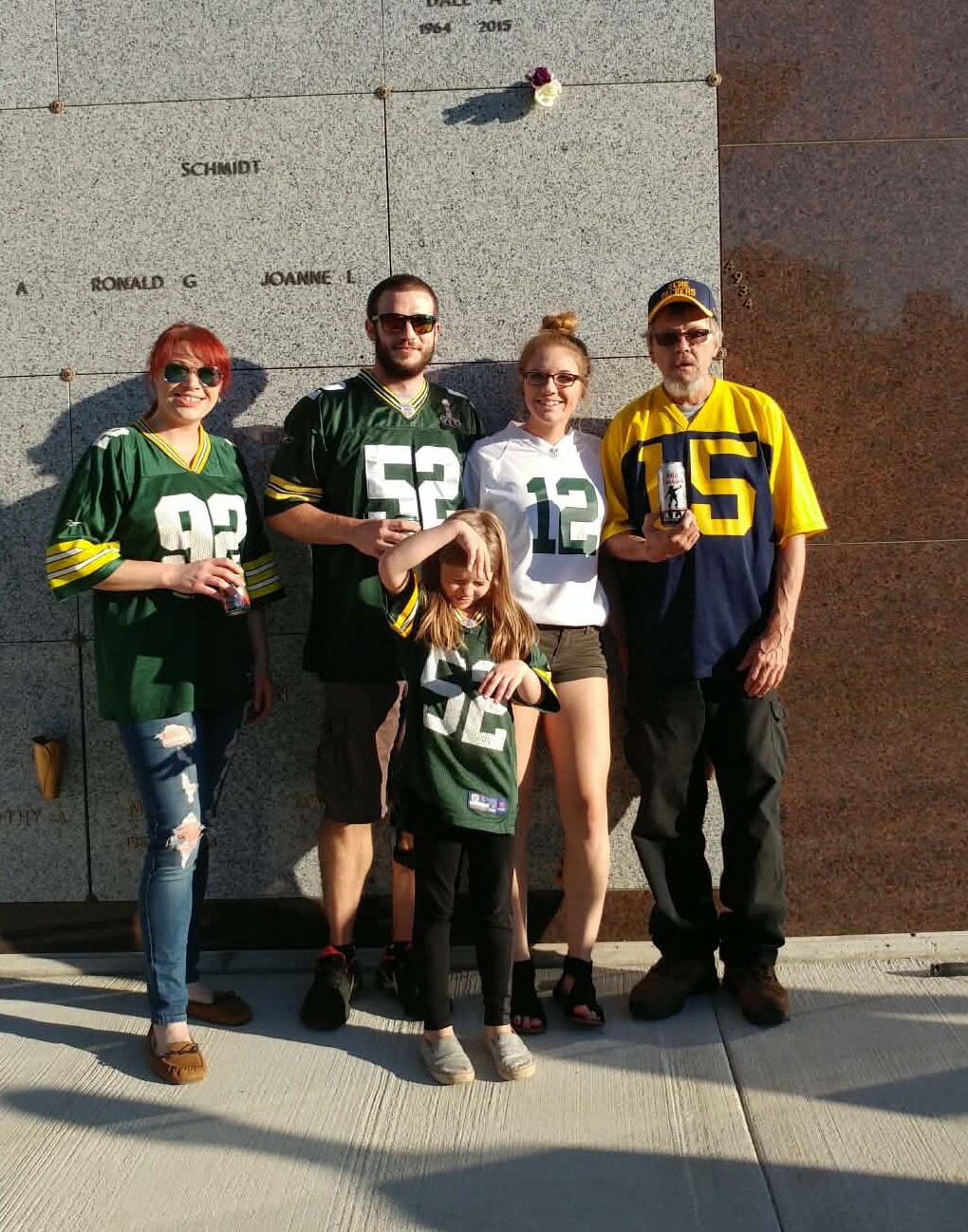 A family of five wearing Green Bay Packers jerseys poses in front of a stone memorial wall with etched names, smiling at the camera.