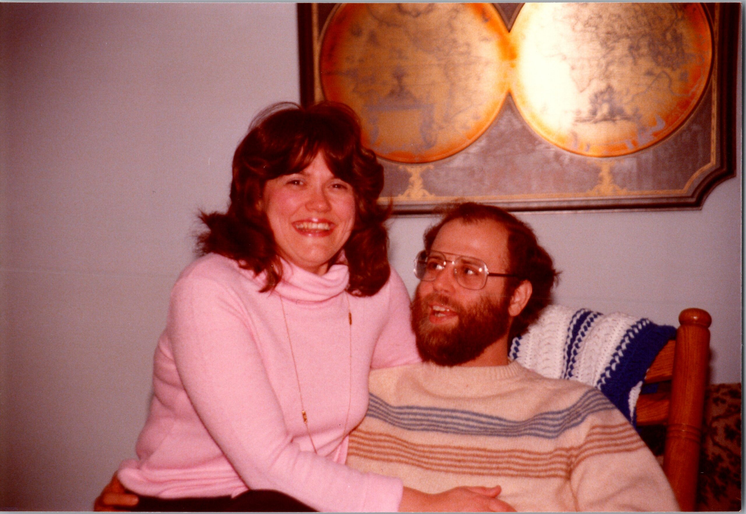 Smiling woman in a pink turtleneck sits beside a bearded man on a bed, a large wall map behind them.