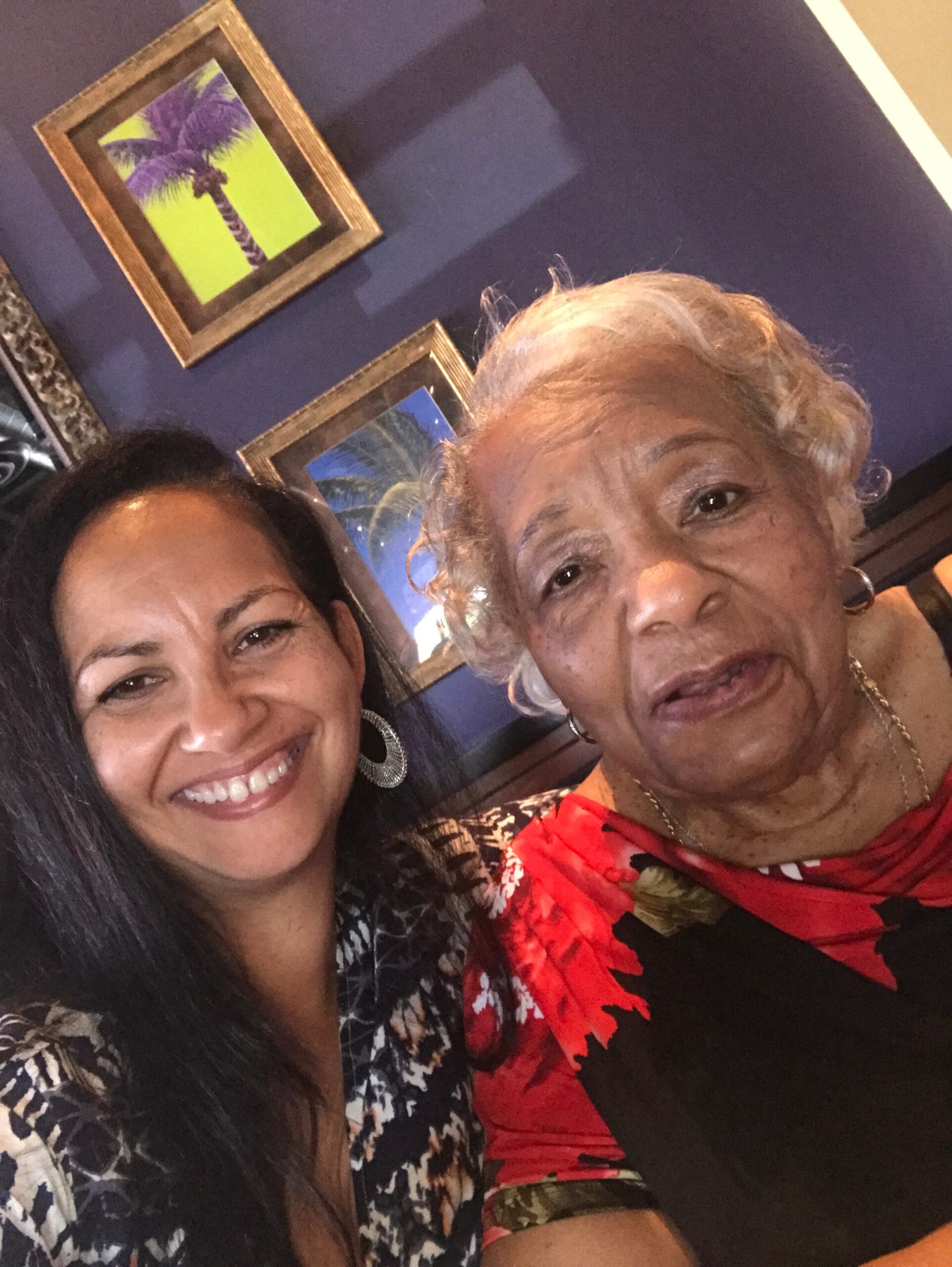 Two women take a close-up selfie indoors, smiling at the camera with blue walls and palm-framed artwork in the background.