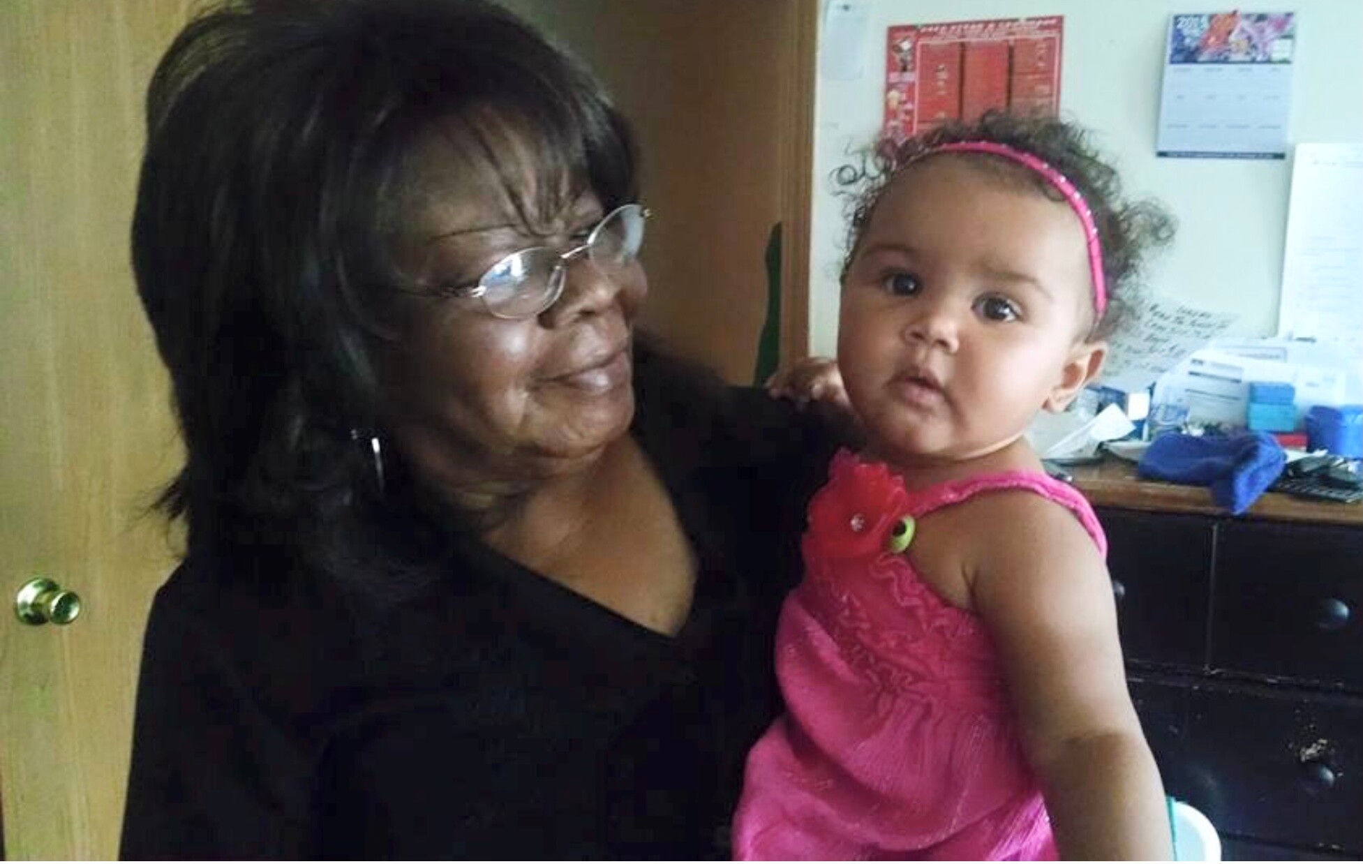 Smiling older woman with glasses holding a baby girl in a pink dress indoors.