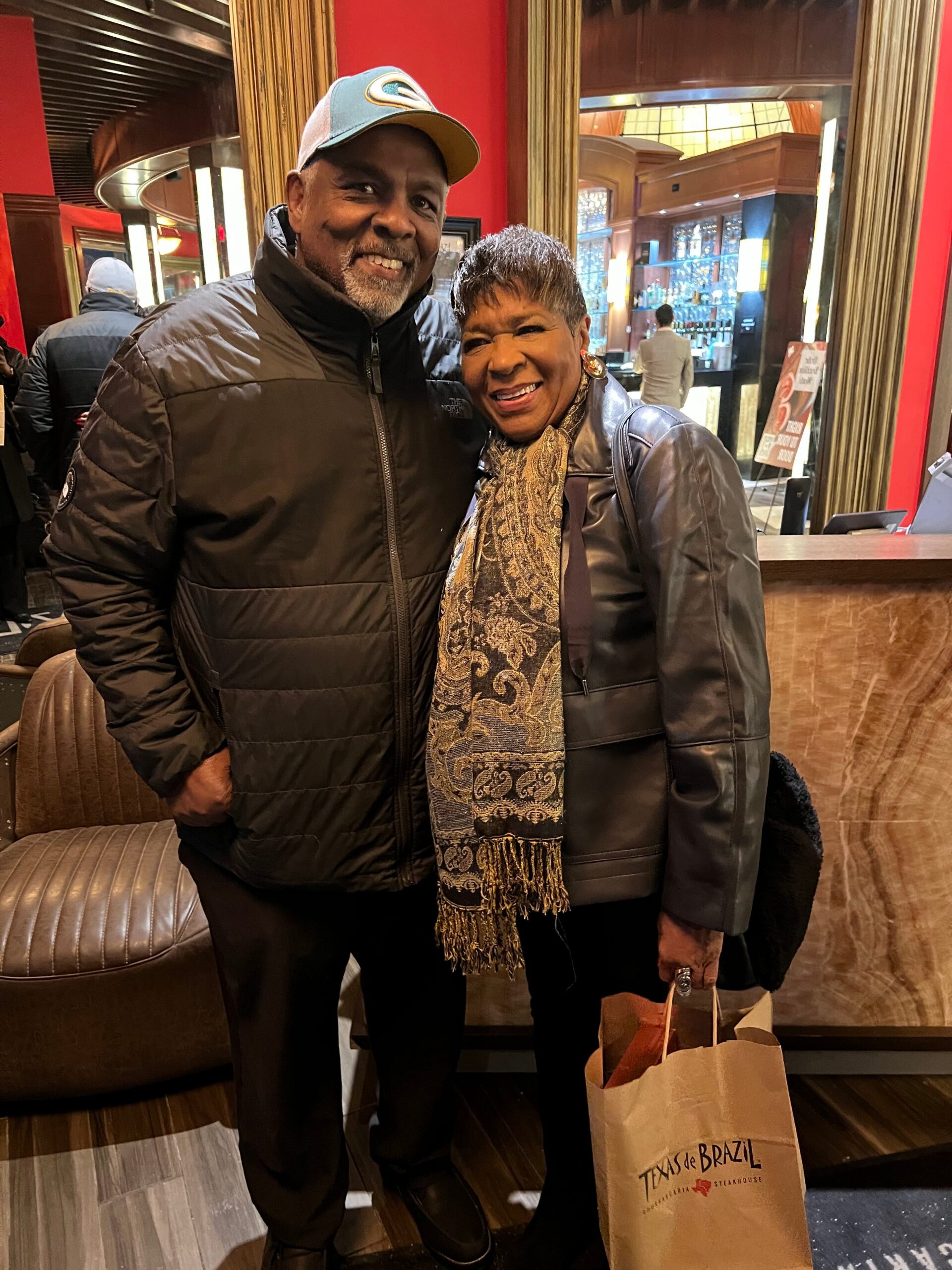 Smiling man and woman posing together in a stylish restaurant lobby; man wears a black jacket and blue cap, woman in a black leather jacket with a patterned scarf, holding a Texas de Brazil bag.