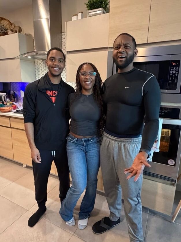 Three people stand together in a modern kitchen, smiling at the camera, with a woman in the middle wearing a gray top and jeans.