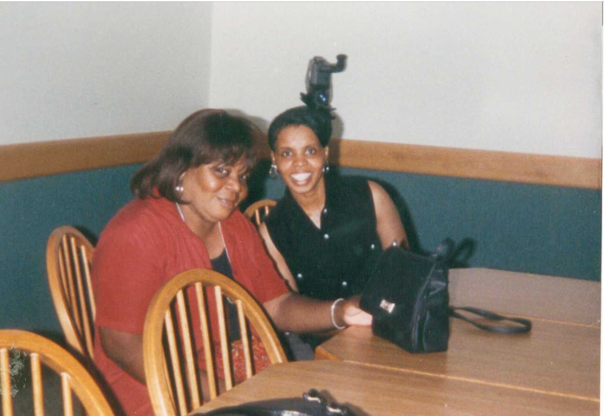 Two women smile while seated at a wooden table in a casual indoor setting; one wears a red top, the other a black sleeveless top with a camera on the table nearby.